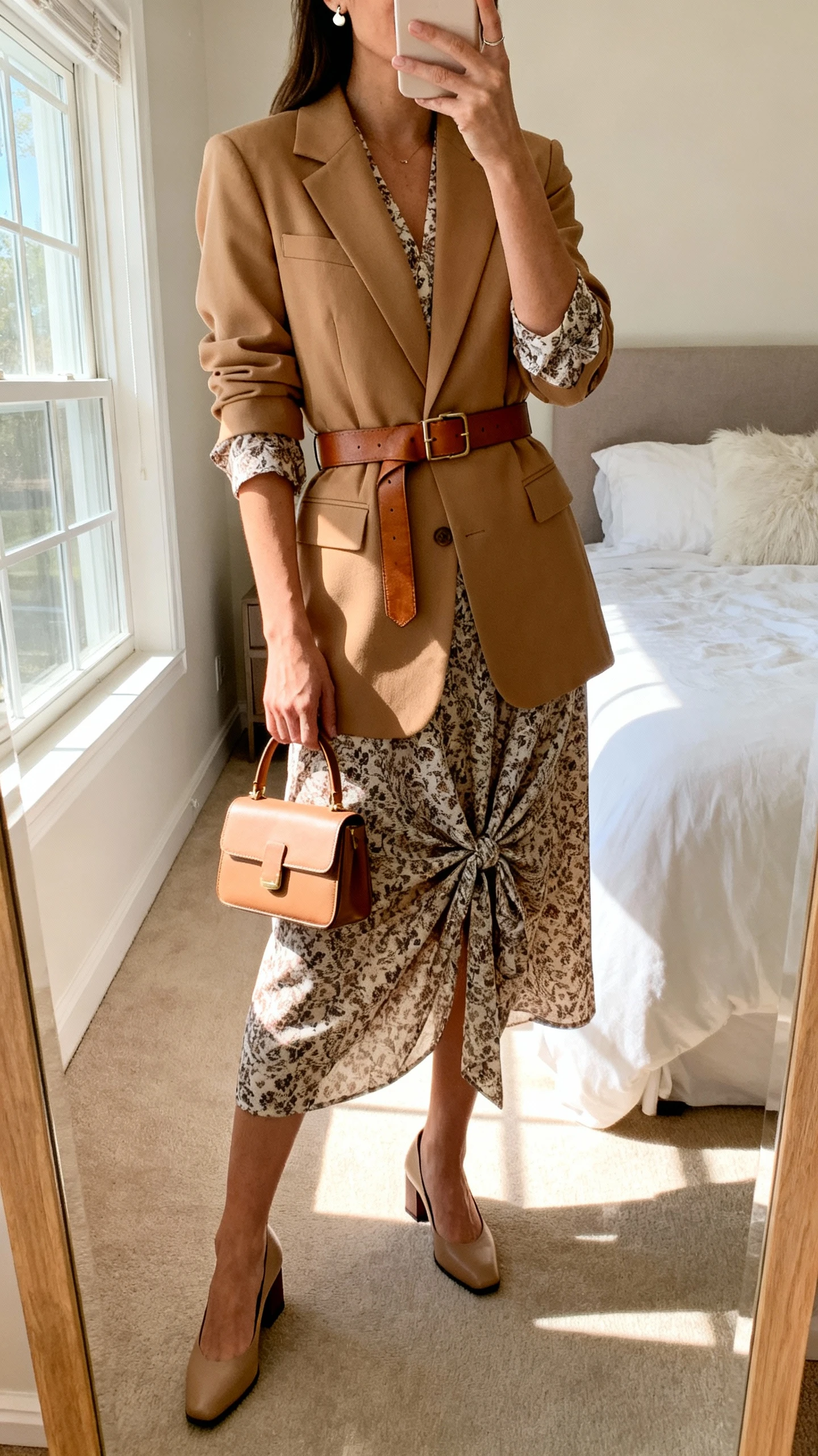 iPhone mirror selfie of a woman wearing a printed midi wrap dress with sleeves, a camel tailored blazer belted at the waist with a medium leather belt, low block-heel pumps, a structured small top-handle bag, and simple stud earrings, face not visible, cropped at neck, bedroom mirror with morning window light, iPhone photo quality.