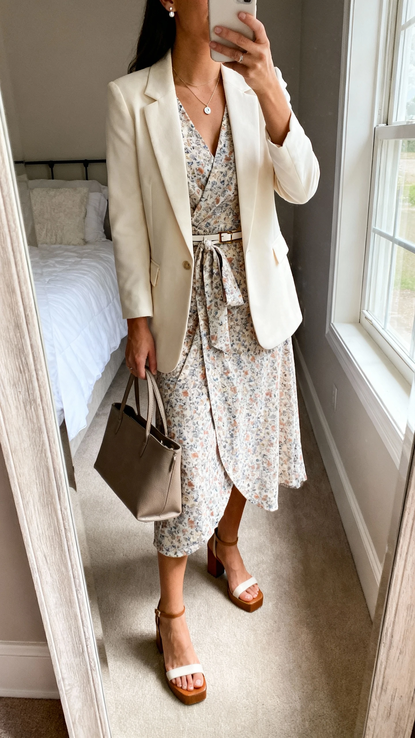 iPhone mirror selfie of a woman wearing a printed midi wrap dress with a cream tailored blazer, block-heel sandals, delicate pendant necklace, simple stud earrings, and a structured tote (optional thin waist belt), face not visible, cropped at neck, bedroom mirror, natural window light, iPhone photo quality.