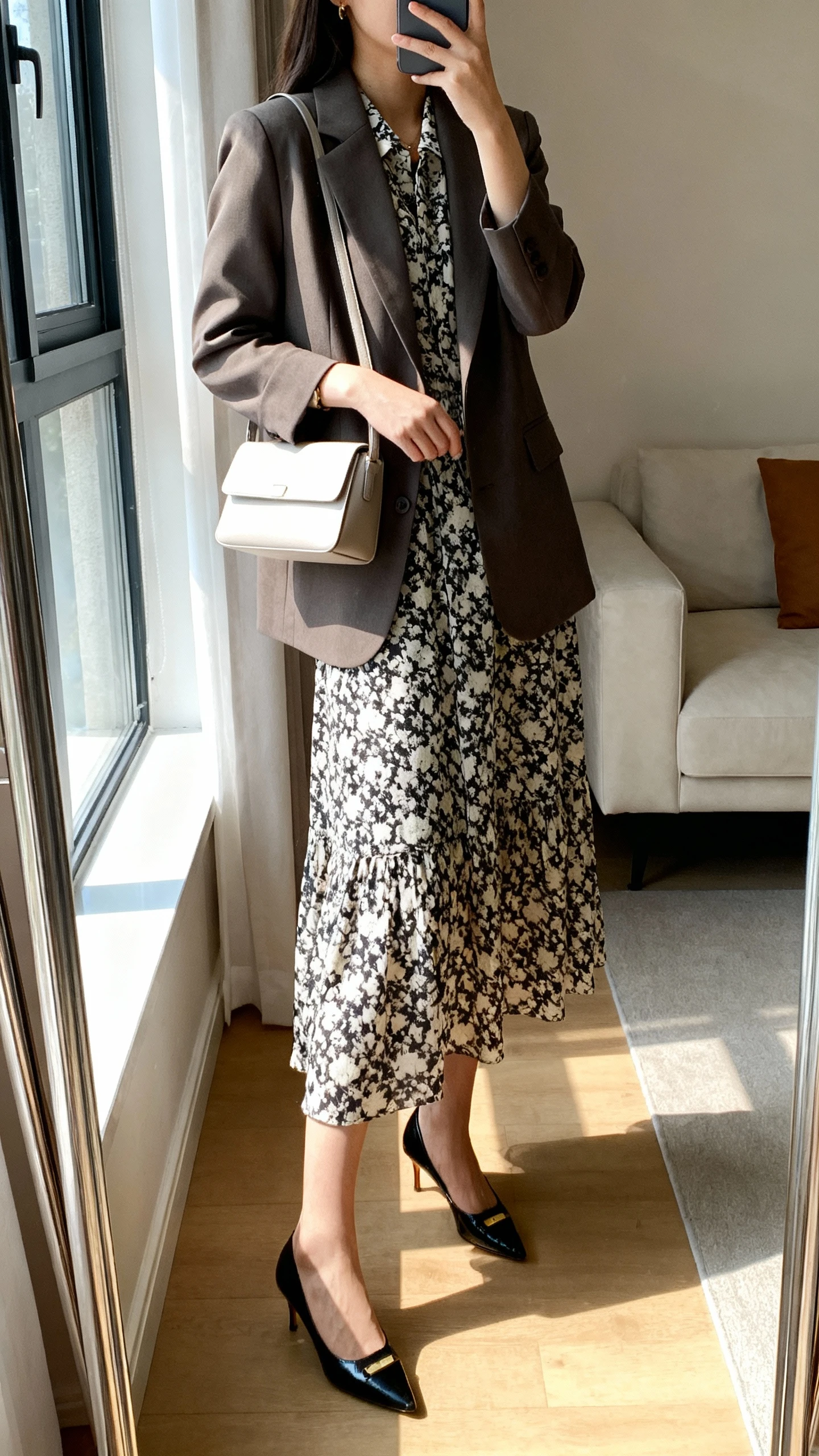iPhone mirror selfie of a woman wearing a printed midi dress layered with a tailored blazer, classic pumps, and a minimalist handbag, face not visible, side angle, living room mirror, natural window light, iPhone photo quality.
