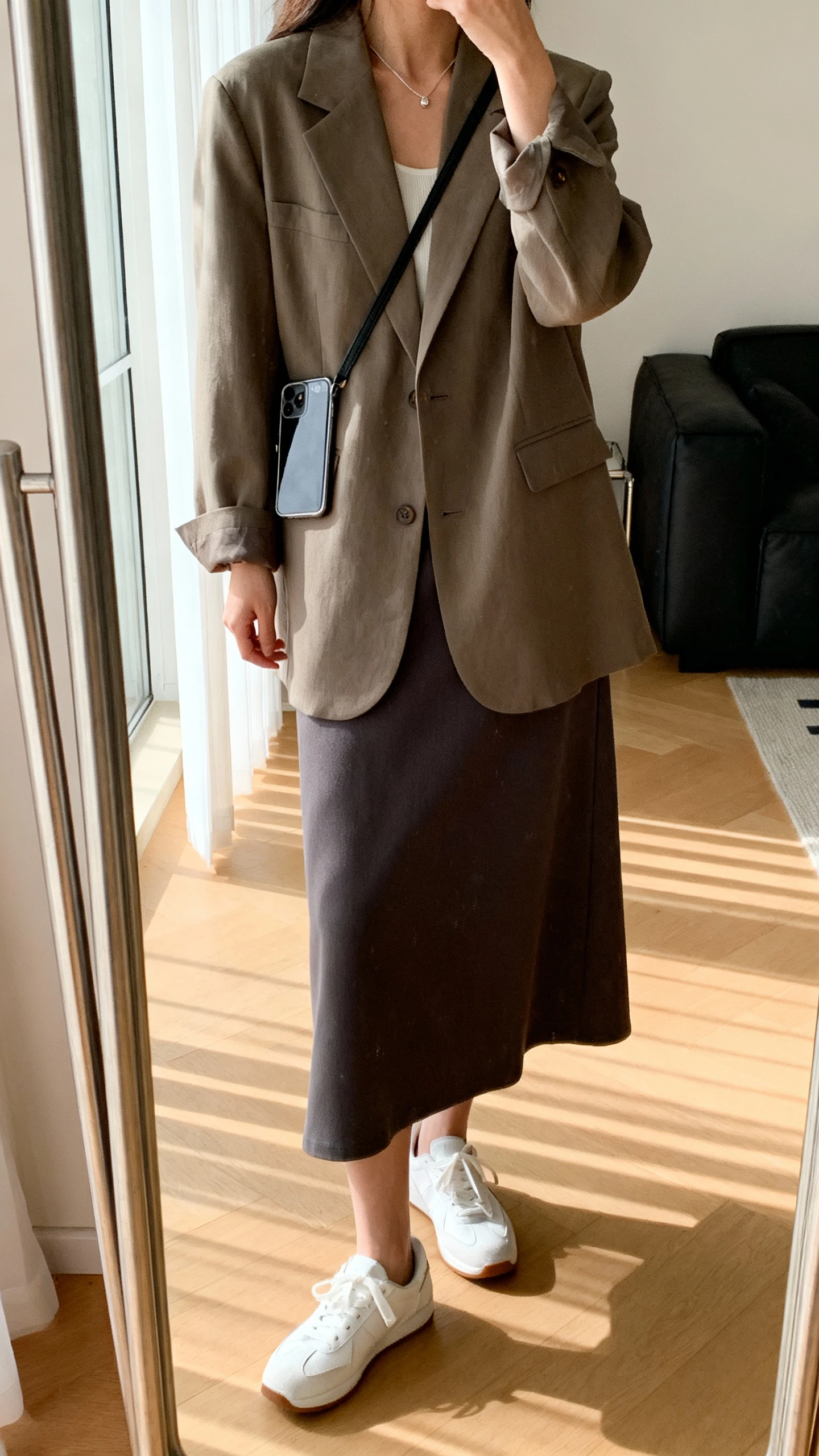 iPhone mirror selfie of a woman wearing a relaxed blazer over a midi skirt with clean white sneakers, minimal necklace, and a crossbody phone bag, face not visible, side angle, living room full-length mirror, indoor natural light, iPhone photo quality.