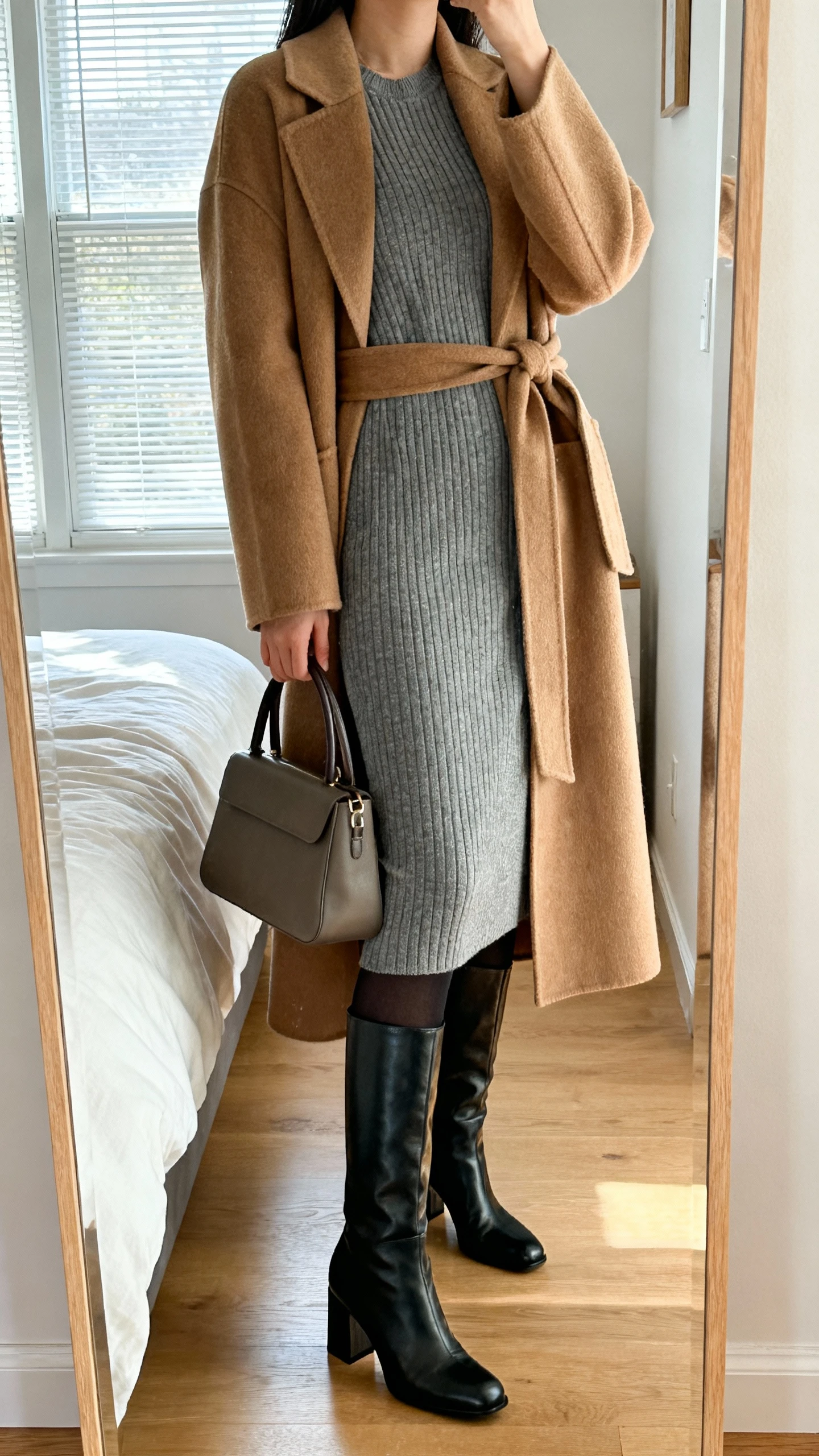 iPhone mirror selfie of a woman wearing a ribbed knit midi dress in heather gray, a camel belted wool wrap coat, black opaque tights, knee-high leather boots with a block heel, and a structured top-handle bag, face not visible, cropped at neck, bedroom mirror with morning window light, iPhone photo quality.
