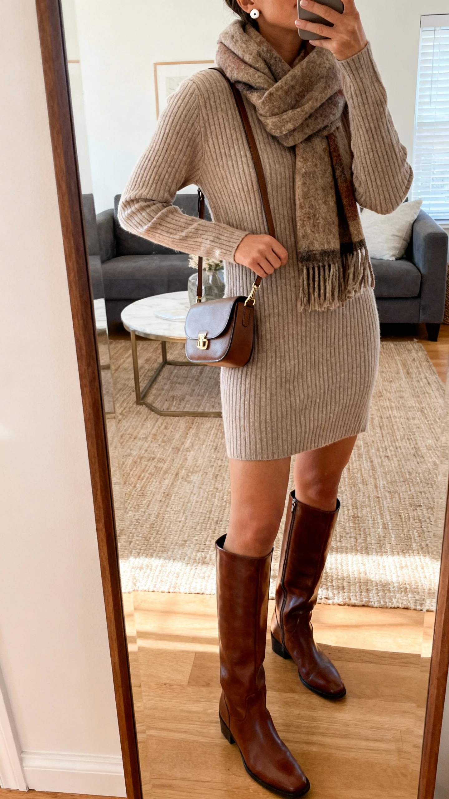 iPhone mirror selfie of a woman wearing a ribbed sweater dress with knee-high leather boots, cozy scarf, small shoulder bag, and stud earrings, face not visible, cropped at neck, living room full-length mirror with warm indoor lighting, iPhone photo quality