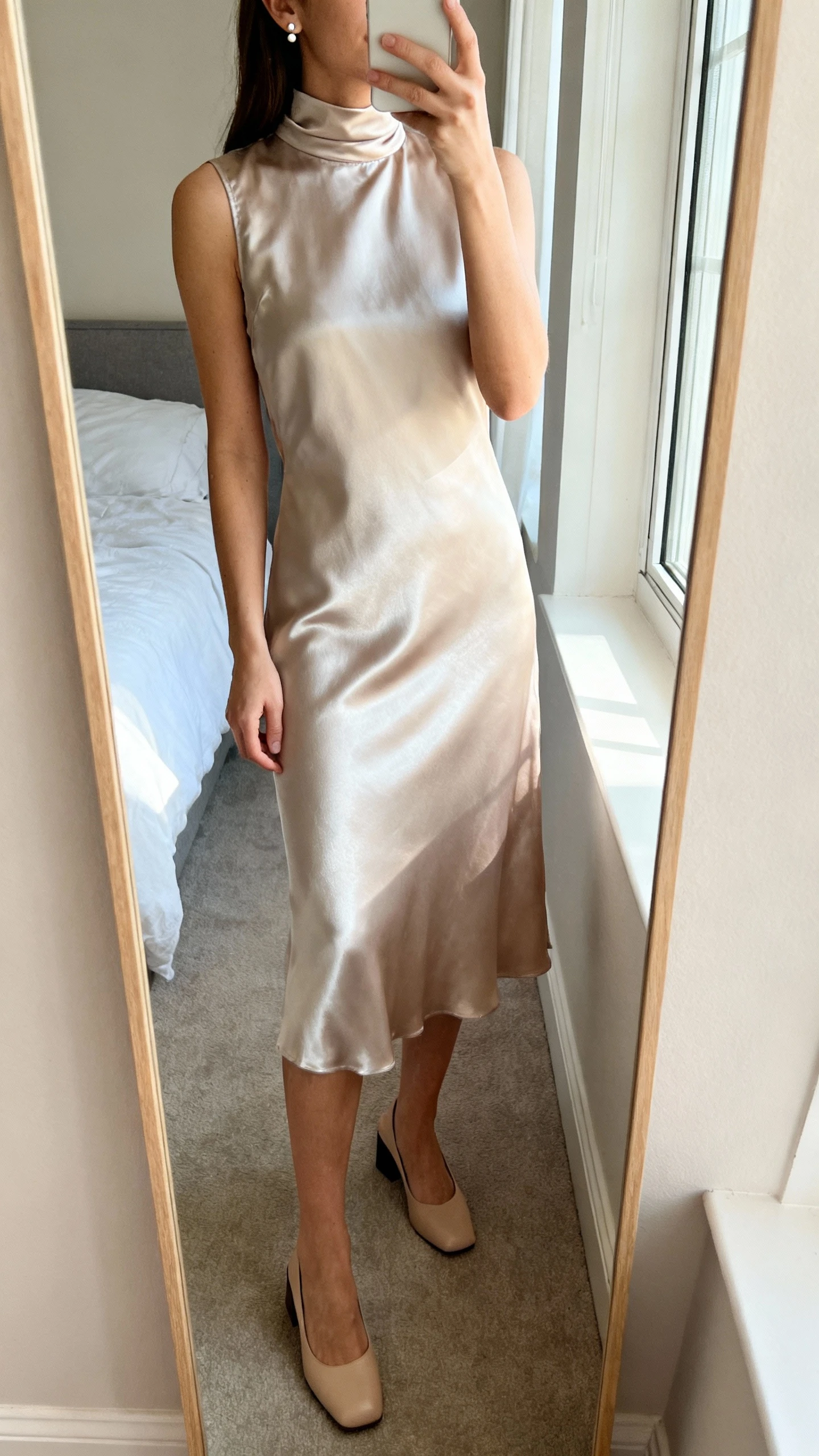 iPhone mirror selfie of a woman wearing a satin high-neck midi dress with a gentle sheen, simple stud earrings and low block heels, face not visible, cropped at neck, bedroom mirror, soft morning window light, iPhone photo quality.