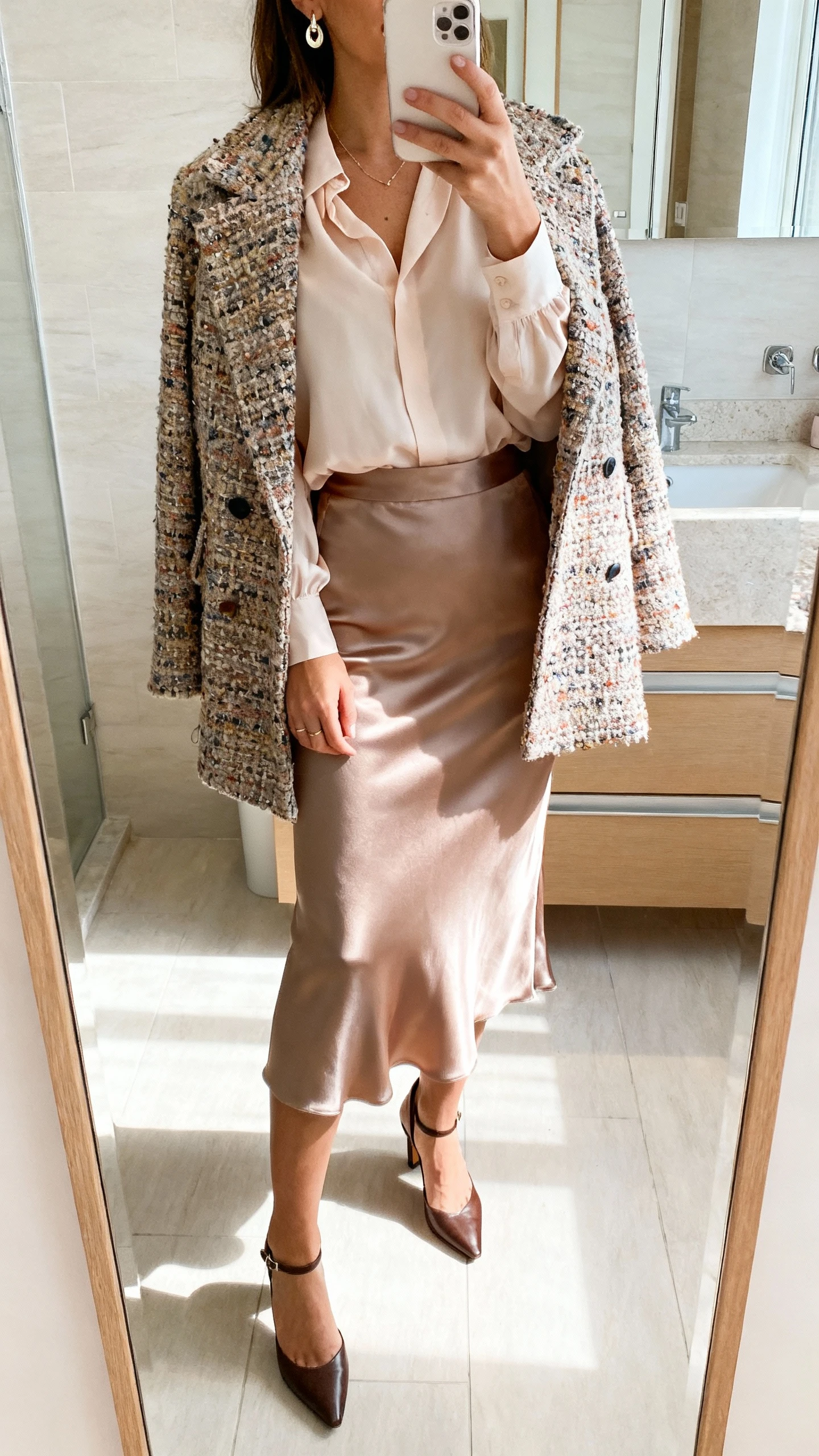 iPhone mirror selfie of a woman wearing a satin midi skirt with a soft blouse and a statement coat, styled with delicate earrings and slingback heels, face not visible, cropped at shoulders, bathroom mirror, bright indoor lighting, iPhone photo quality.