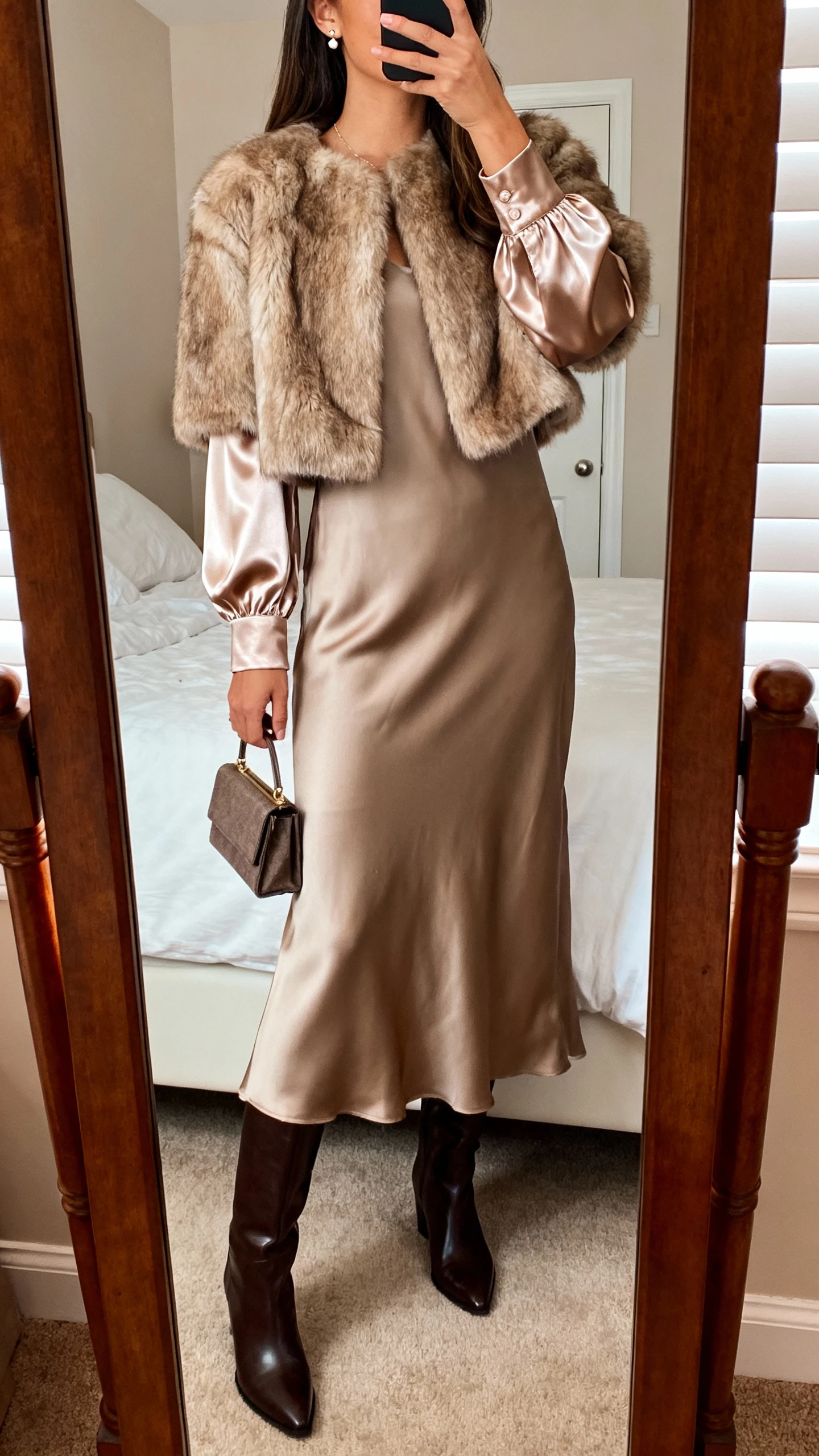 iPhone mirror selfie of a woman wearing a satin-sleeved crepe maxi dress under a faux-fur short jacket, sleek knee-high boots, small structured clutch, and delicate stud earrings, face not visible, cropped at shoulders, bedroom mirror, warm indoor lighting, iPhone photo quality.
