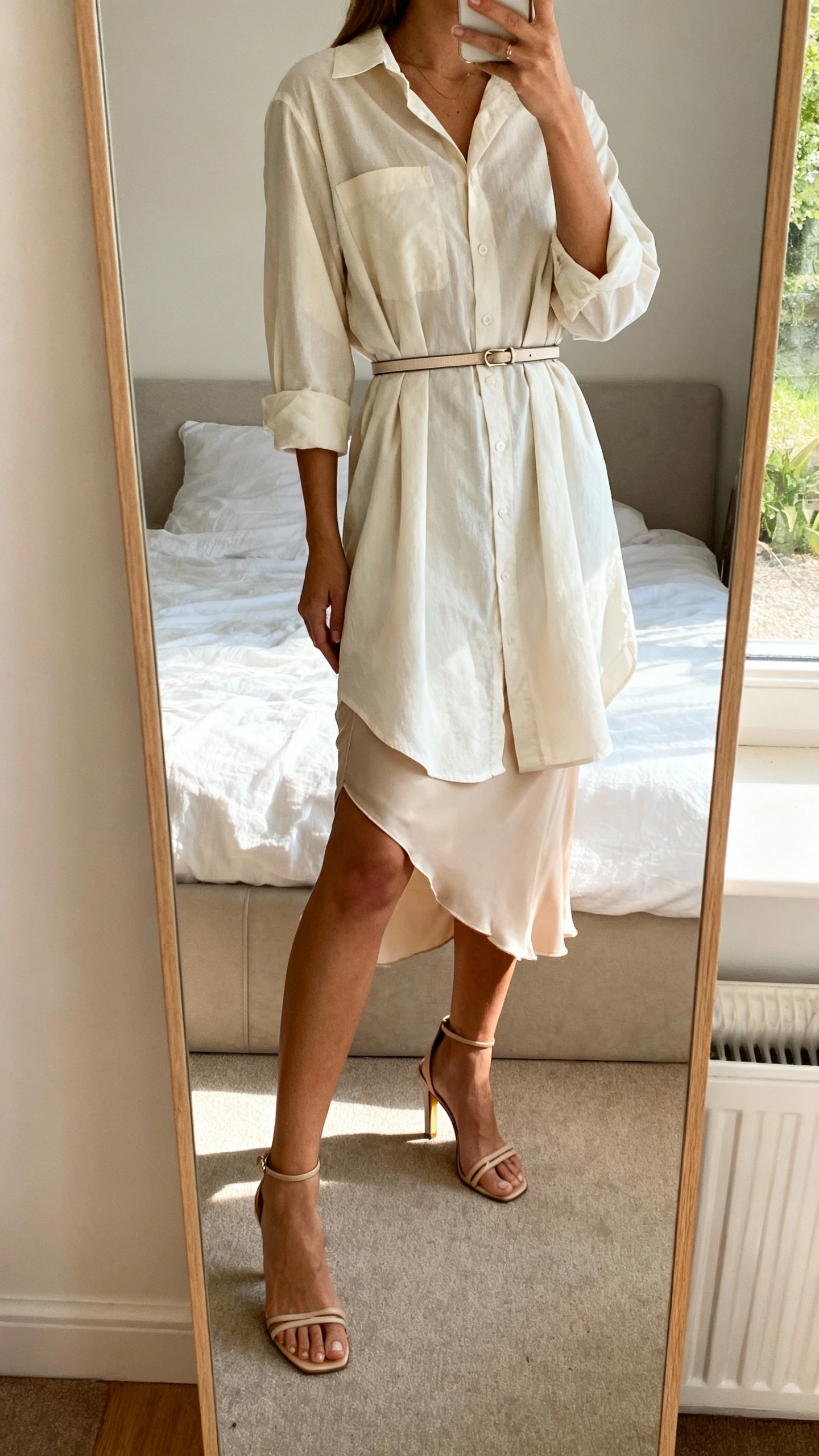 iPhone mirror selfie of a woman wearing a shirt dress layered over a slip skirt peeking below the hem, with heeled sandals and a slim belt; face not visible, cropped at shoulders, bedroom mirror, natural daylight, iPhone photo quality.