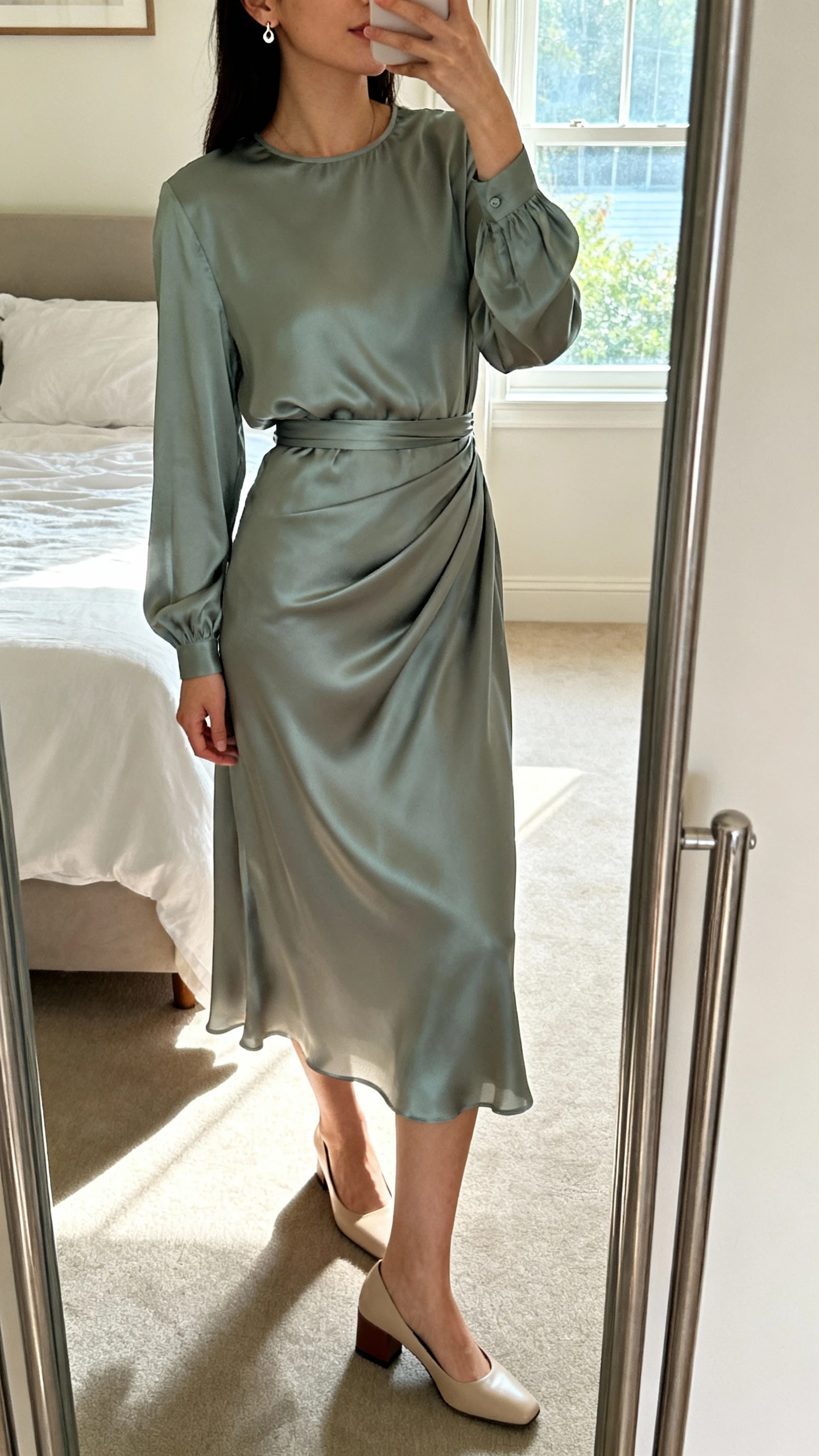 iPhone mirror selfie of a woman wearing a silk-soft long-sleeve midi dress that drapes and floats with a secure waist, paired with delicate stud earrings and low block-heel pumps, face not visible, cropped at neck, bedroom mirror, morning window light, iPhone photo quality.