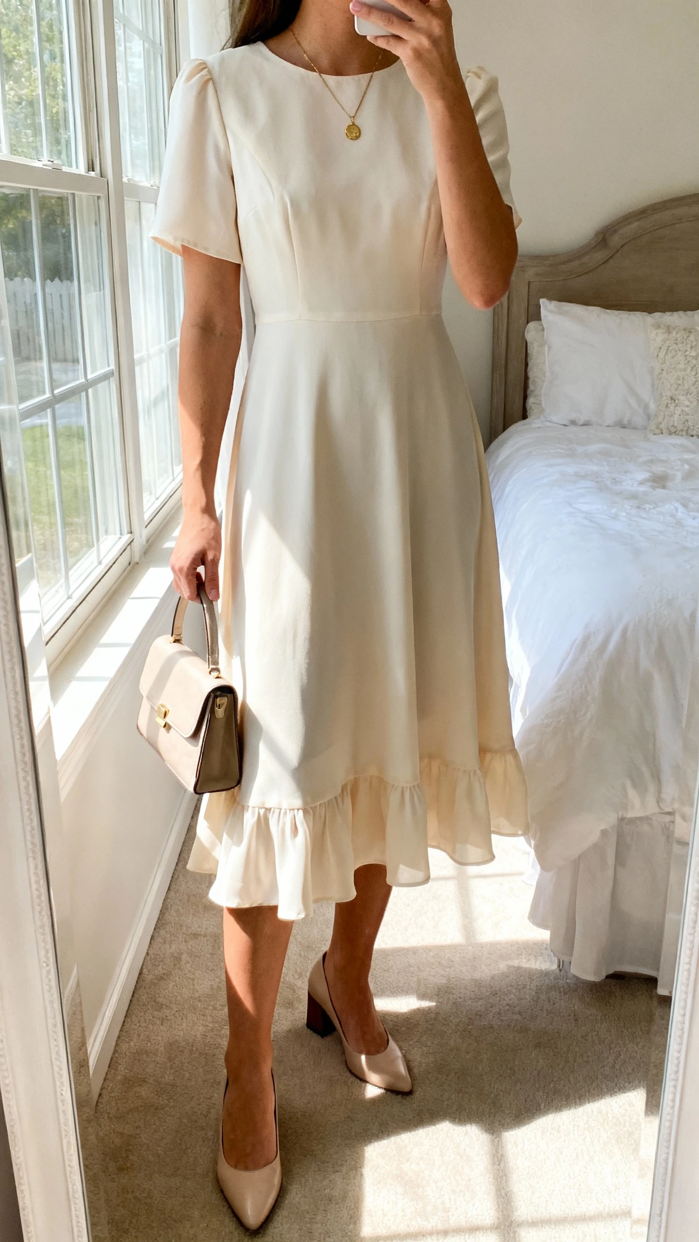 iPhone mirror selfie of a woman wearing a soft A-line church dress with gentle movement, midi length, subtle flutter hem, delicate pendant necklace, small structured handbag, and low block-heel pumps; face not visible, cropped at neck, bedroom mirror with natural window light, iPhone photo quality.