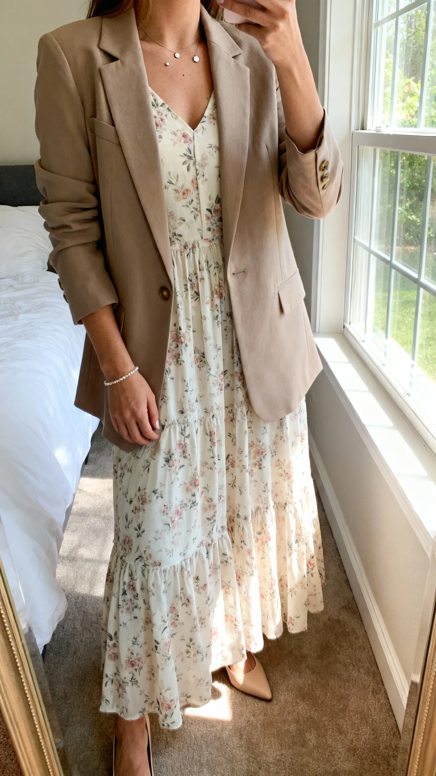 iPhone mirror selfie of a woman wearing a soft floral maxi dress with a tailored blazer, delicate studs, simple bracelet, and classic low heels, face not visible, cropped at neck, bedroom mirror, natural window light, iPhone photo quality