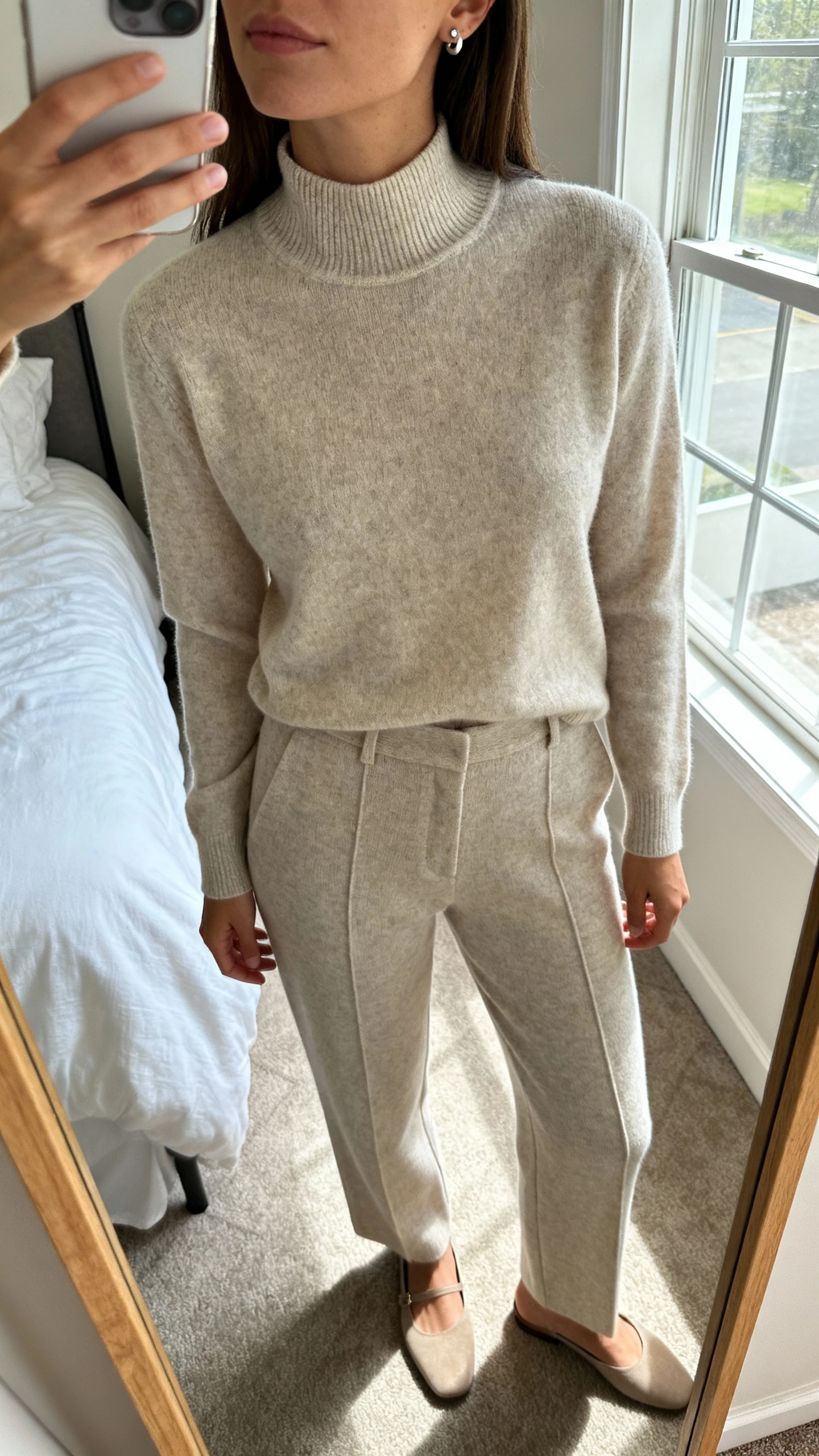 iPhone mirror selfie of a woman wearing a soft knit mock-neck top with matching knit pants set that looks tailored, plus minimal stud earrings and simple flats, face not visible, cropped at neck, bedroom mirror, natural window light, iPhone photo quality