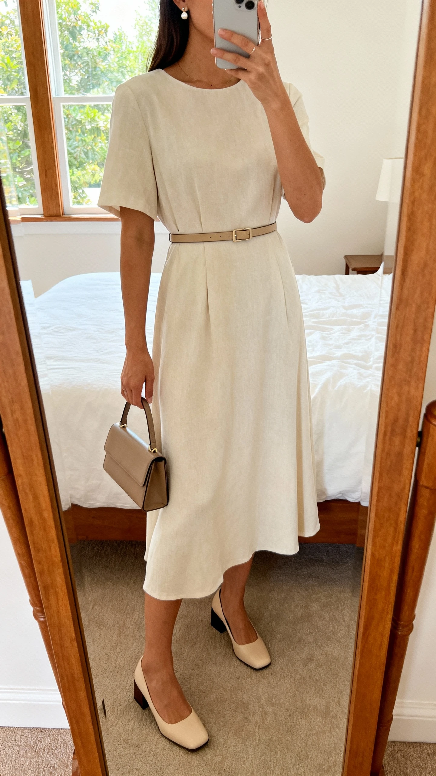 iPhone mirror selfie of a woman wearing a soft midi dress cinched with a slim belt, low block heels, delicate stud earrings, and a small structured handbag, face not visible, cropped at neck, bedroom mirror, natural window light, iPhone photo quality.