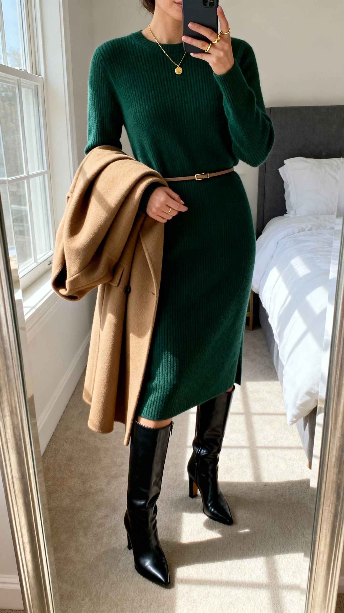 iPhone mirror selfie of a woman wearing a soft midi sweater dress (ribbed or smooth) in forest green with a thin waist belt, knee-high heeled boots in black leather, a tailored camel wool coat draped over arm, delicate gold necklace and ring, face not visible, cropped at neck, bedroom full-length mirror, natural morning window light, iPhone photo quality.