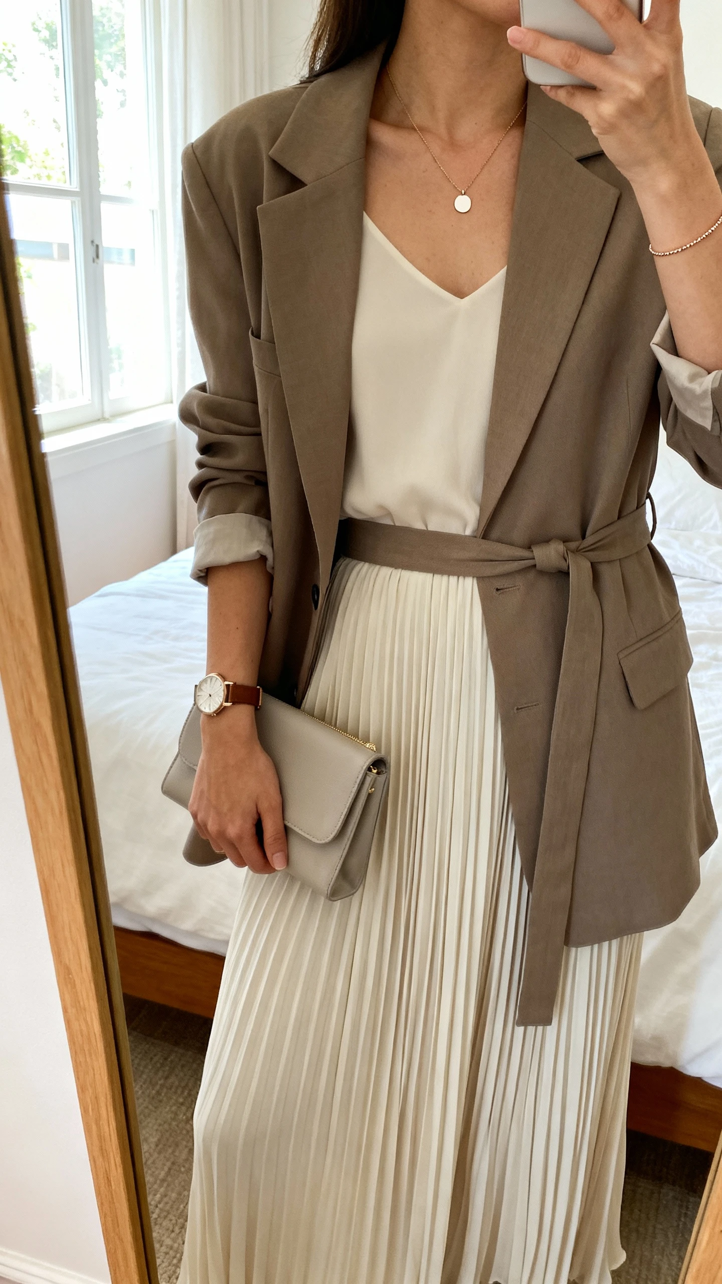 iPhone mirror selfie of a woman wearing a soft pleated maxi dress with a belted blazer, minimal pendant necklace, delicate watch, and simple clutch, face not visible, cropped at neck, bedroom mirror, natural window light, iPhone photo quality