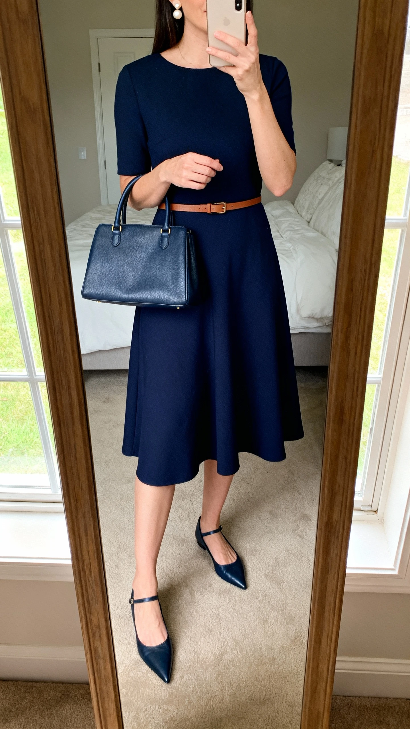 iPhone mirror selfie of a woman wearing a solid navy soft A-line midi dress with a thin leather belt, pointed-toe flats, structured top-handle bag, and pearl stud earrings, face not visible, cropped at neck, bedroom mirror, natural window light, iPhone photo quality.