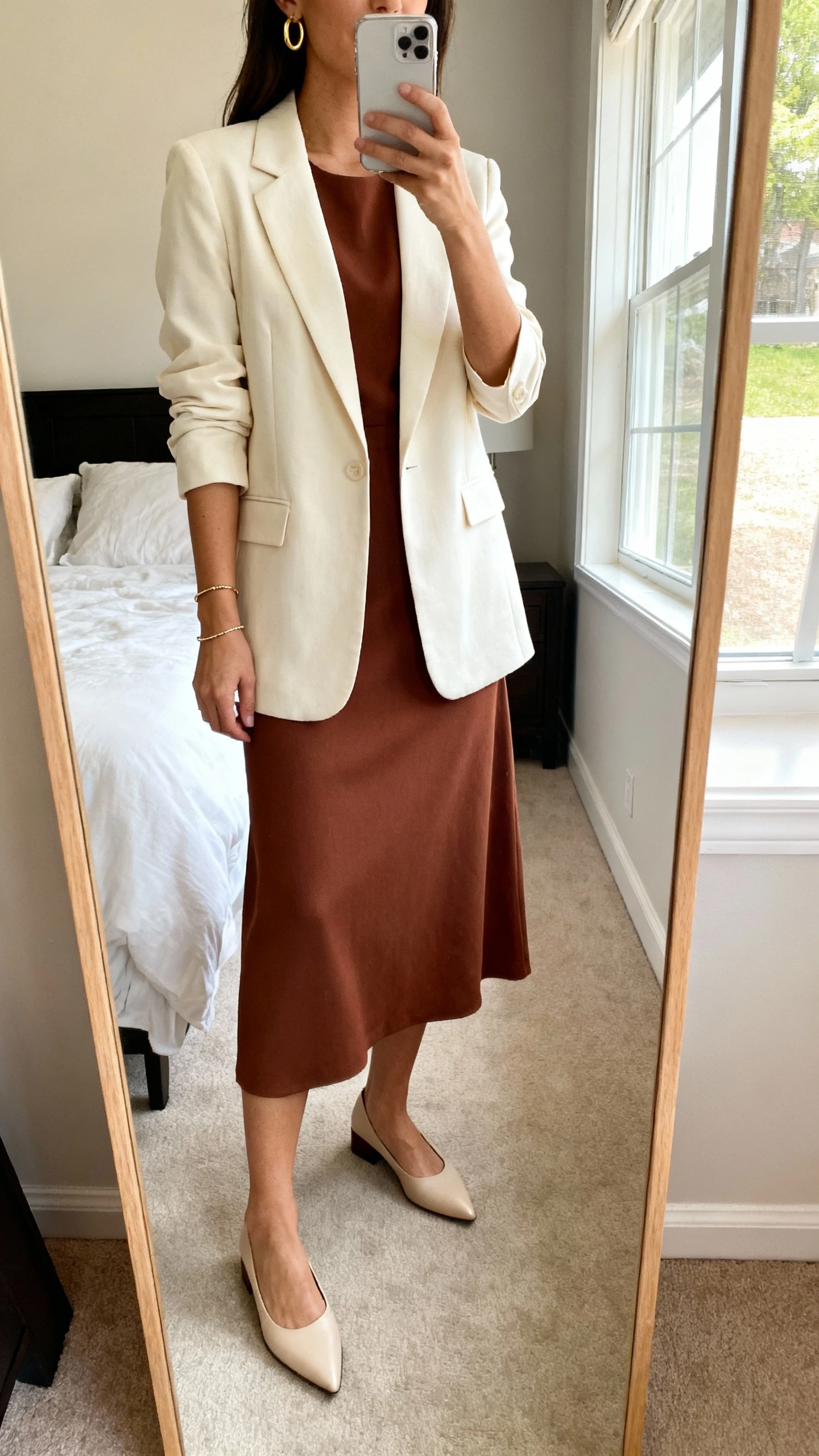 iPhone mirror selfie of a woman wearing a structured solid-color midi dress with bracelet-length sleeves, a single-breasted cream blazer, pointed-toe low block heels, simple gold hoop earrings, and a delicate bracelet, face not visible, cropped at neck, bedroom mirror with natural window light, iPhone photo quality.