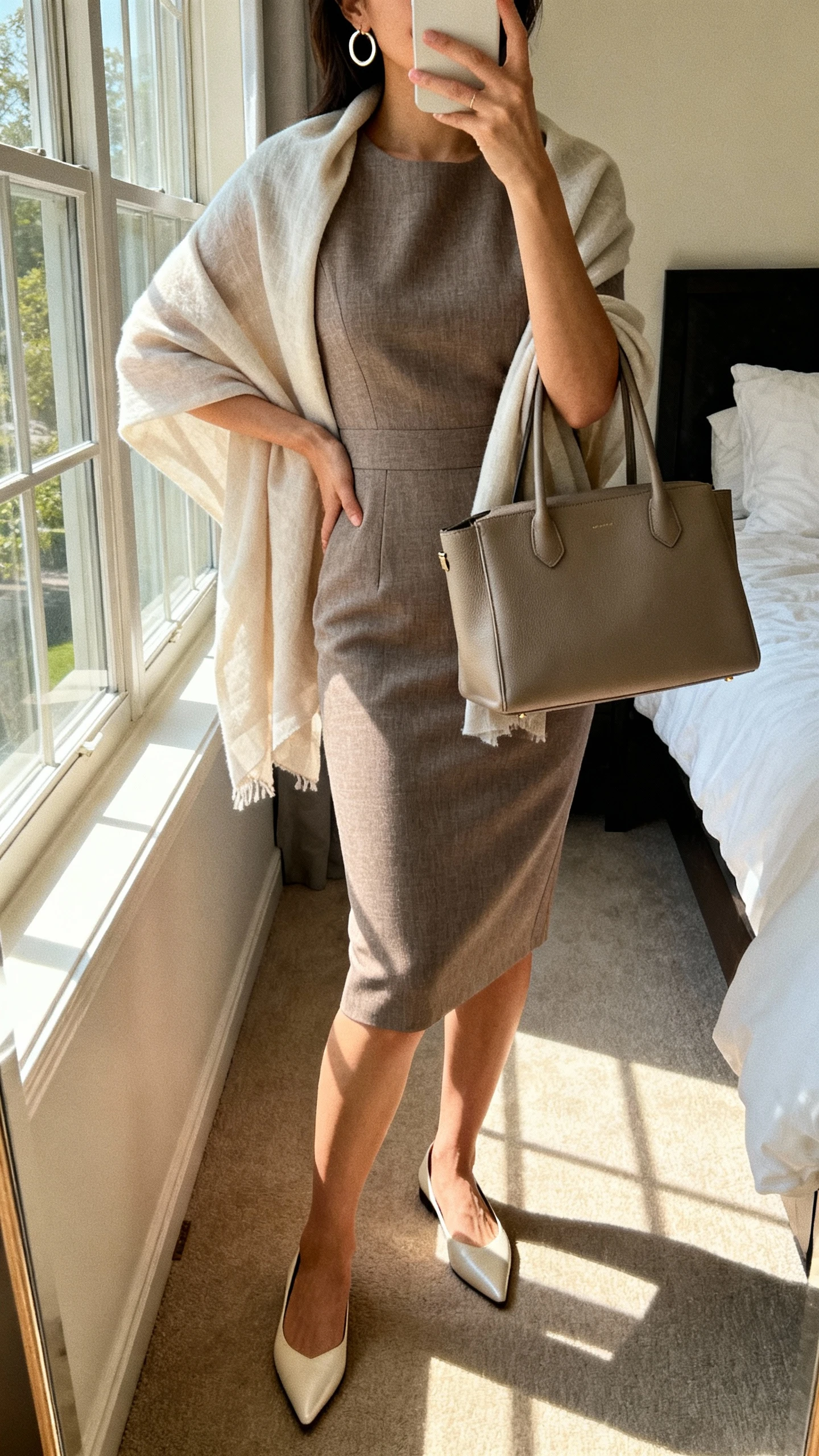 iPhone mirror selfie of a woman wearing a tailored sheath dress with a soft layer (lightweight shawl or chiffon overlay), minimal hoop earrings, structured tote, and pointed-toe flats; face not visible, cropped at shoulders, bedroom mirror, natural window light, iPhone photo quality.
