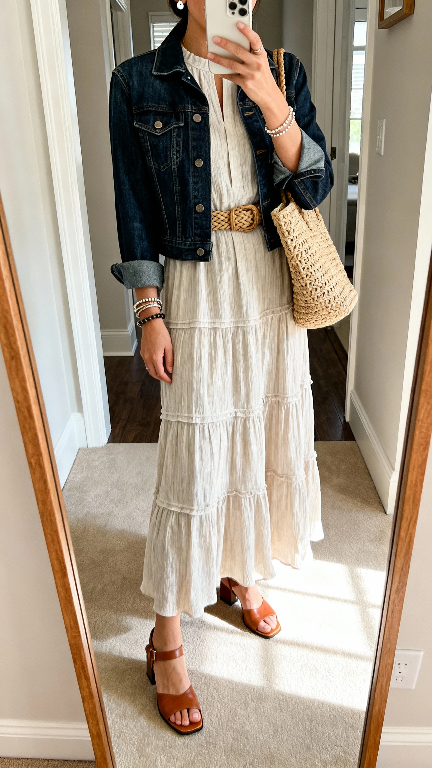 iPhone mirror selfie of a woman wearing a tiered maxi dress with sleeves and modest neckline, a slightly structured dark-wash denim jacket, low block-heel leather sandals, an optional woven belt, stacked bracelets, simple stud earrings, and a woven tote; face not visible, cropped at neck, entryway mirror with natural light, iPhone photo quality.