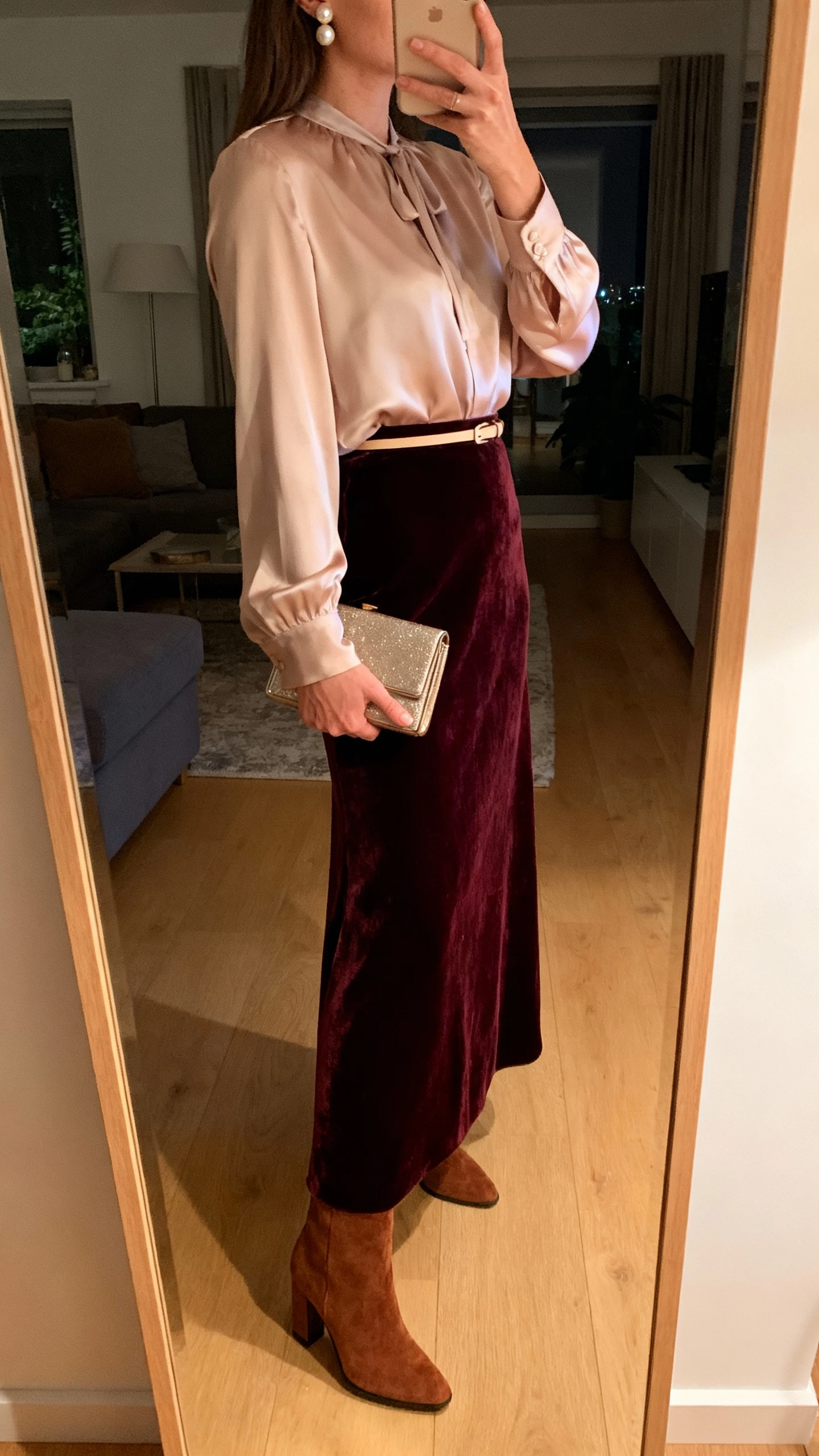 iPhone mirror selfie of a woman wearing a velvet maxi skirt with a soft silk‑blend blouse, thin waist belt, suede heeled boots, pearl studs, and a compact clutch; face not visible, side angle, living room full‑length mirror, evening indoor lighting, iPhone photo quality.