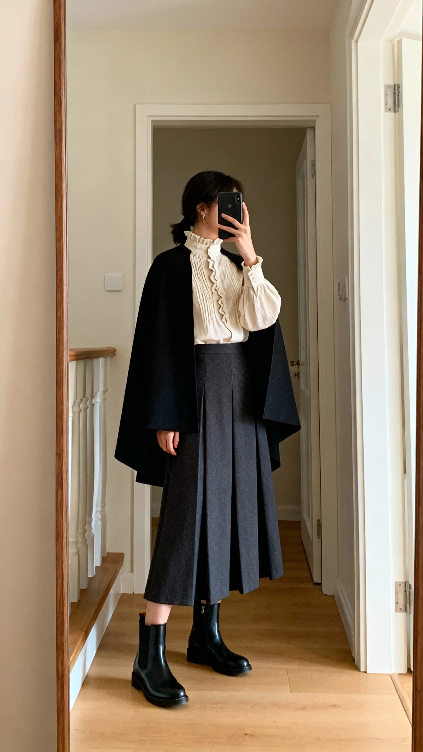 iPhone mirror selfie of a woman wearing a Victorian-inspired high-neck blouse with an A-line maxi skirt, sleek Chelsea boots, and a structured cape, face not visible, side angle, entryway mirror, warm indoor lighting, iPhone photo quality.