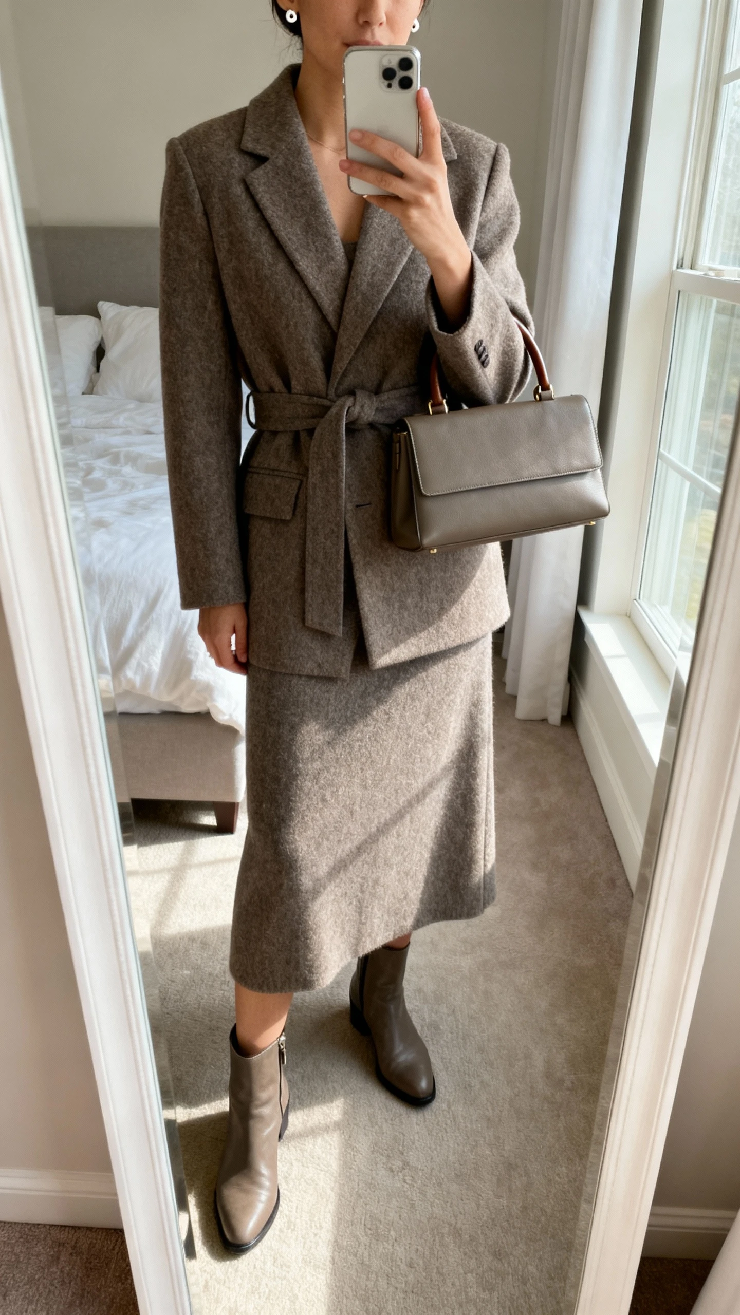 iPhone mirror selfie of a woman wearing a wool midi dress with a belted, tailored wool coat, simple leather ankle boots, structured top-handle bag, and minimal stud earrings, face not visible, cropped at neck, bedroom mirror, soft morning window light, iPhone photo quality.