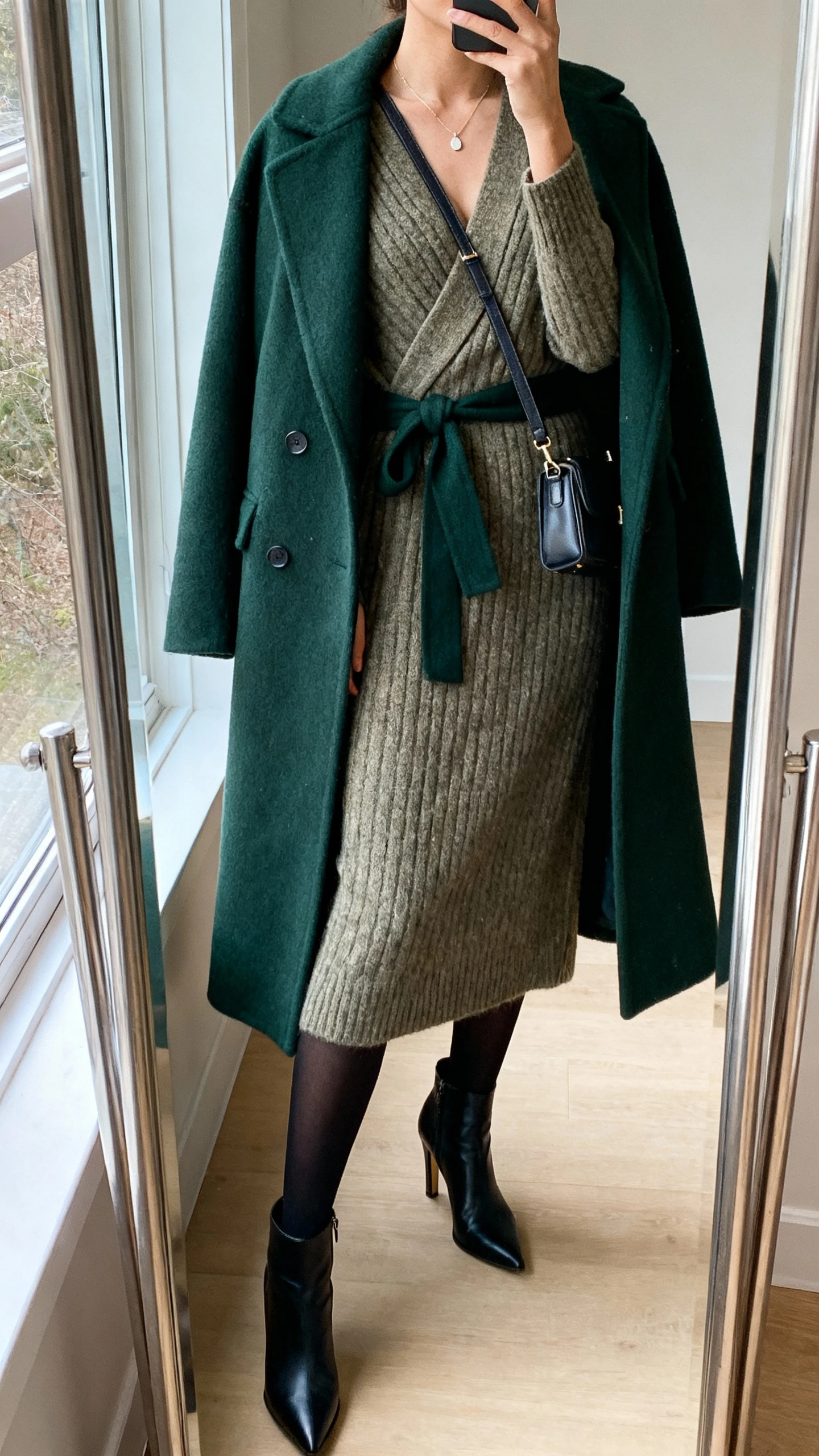 iPhone mirror selfie of a woman wearing a wrap-style knit midi dress, a belted wool coat in deep green, heeled ankle boots, black opaque tights, a minimal pendant necklace, and a structured crossbody; face not visible, cropped at neck, full-length mirror by window, natural morning light, iPhone photo quality.