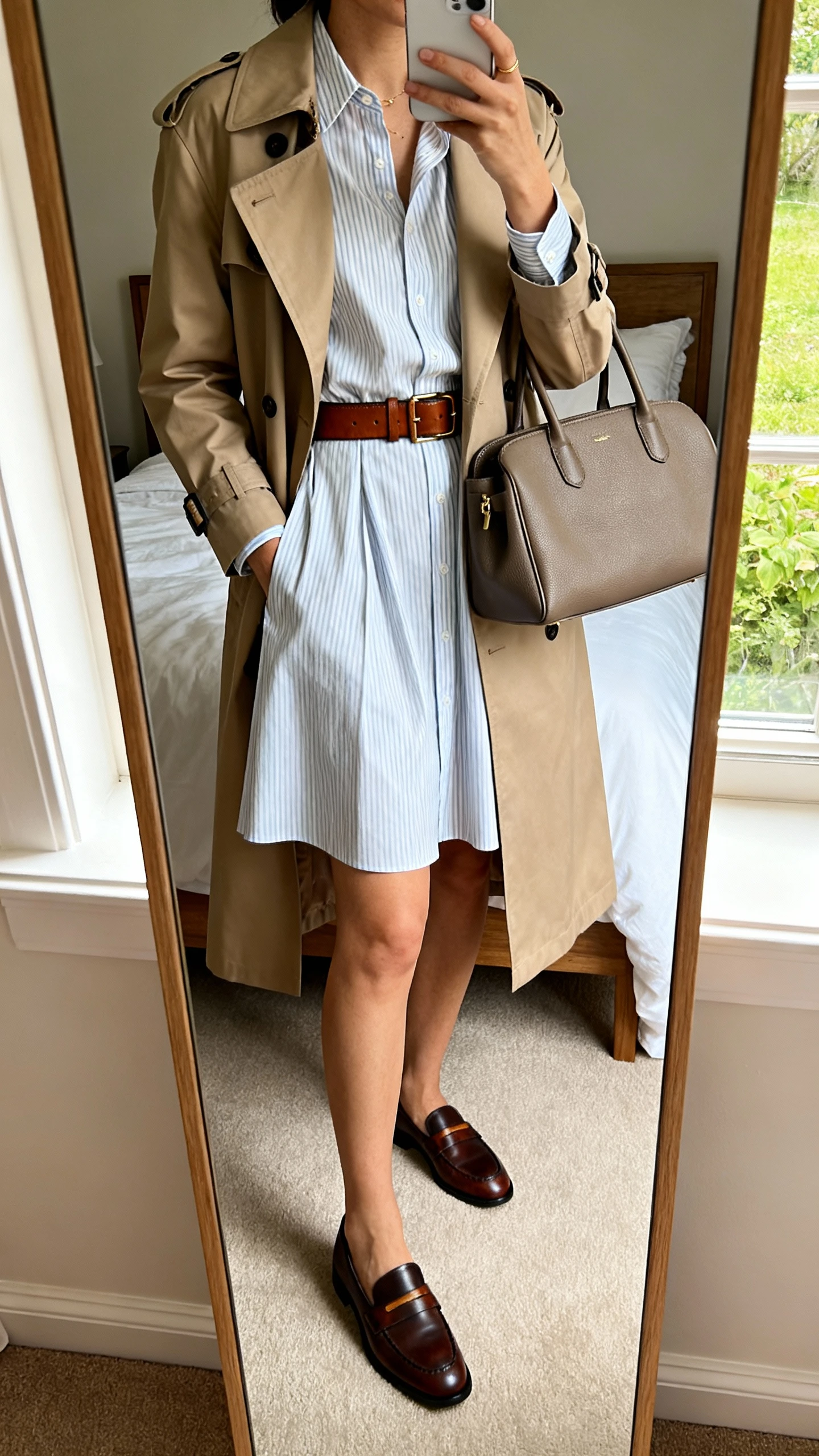 iPhone mirror selfie of a woman wearing an A-line shirt dress layered with a trench coat, leather belt, loafers, and a structured tote, face not visible, cropped at shoulders, bedroom mirror, natural daylight from window, iPhone photo quality.