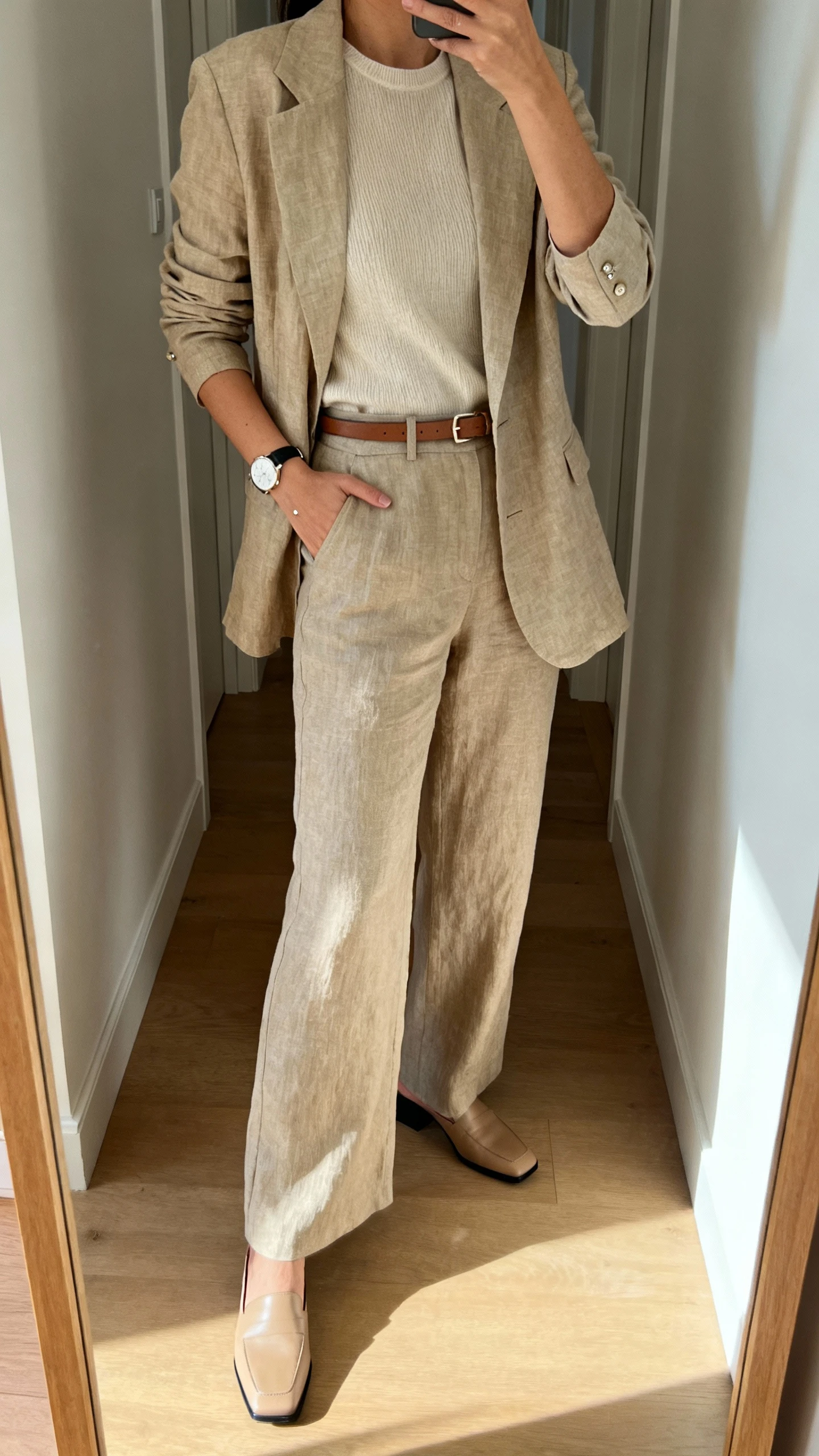 iPhone mirror selfie of a woman wearing an unlined or half-lined linen-blend or tropical-wool blazer over an elbow-sleeve knit top with a crew neckline, high-waisted wide-leg trousers in a matching or complementary tone, low block-heel mules or loafers, a thin leather belt, classic watch, and subtle studs; face not visible, cropped at shoulders, full-length mirror in hallway, indoor daylight, iPhone photo quality.
