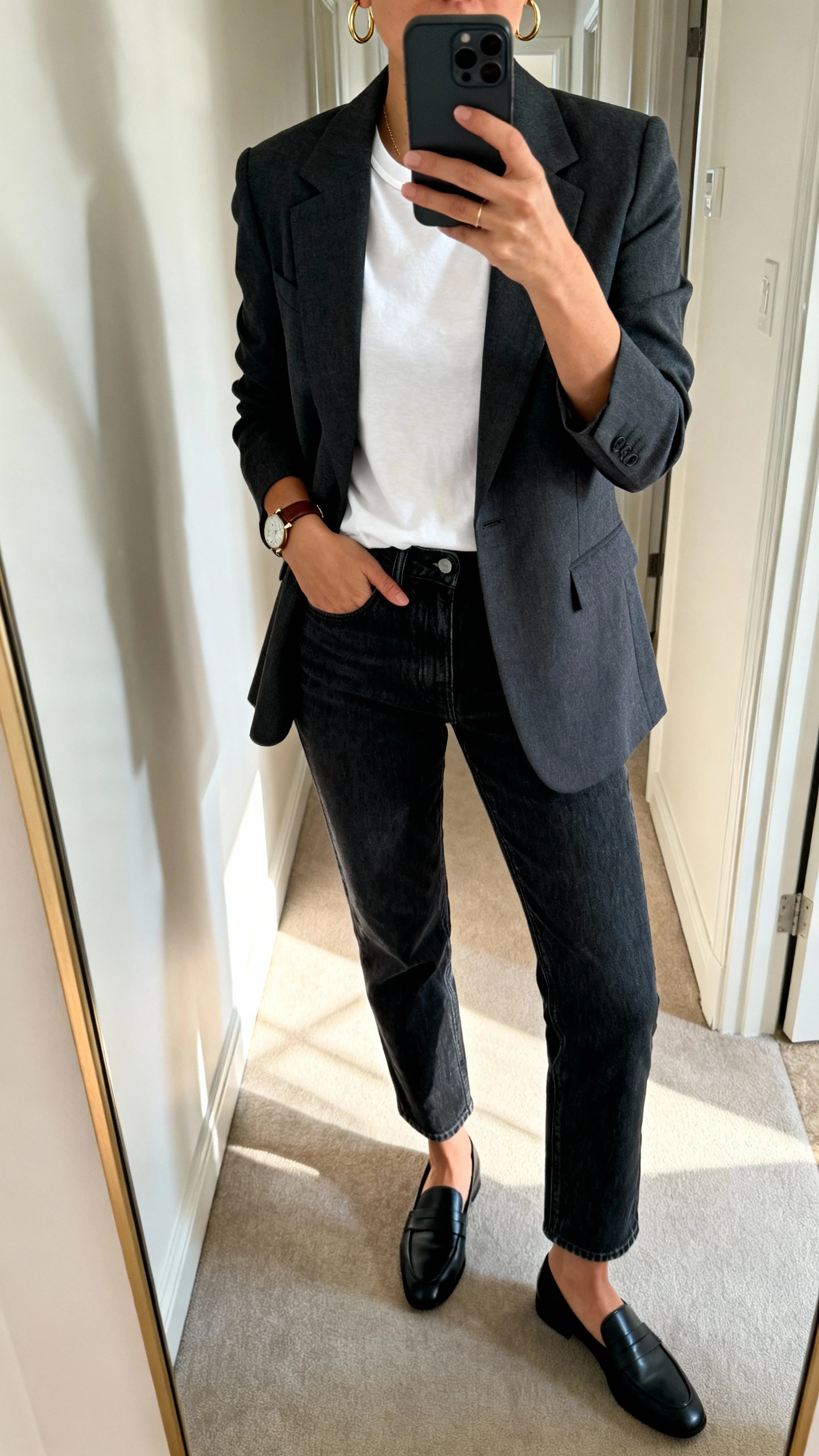 iPhone mirror selfie of a woman wearing dark-wash straight-leg denim, a tailored charcoal blazer over a white crew tee, black loafers, delicate gold hoop earrings, and a leather watch, face not visible, cropped at shoulders, full-length mirror in hallway with indoor daylight, iPhone photo quality.