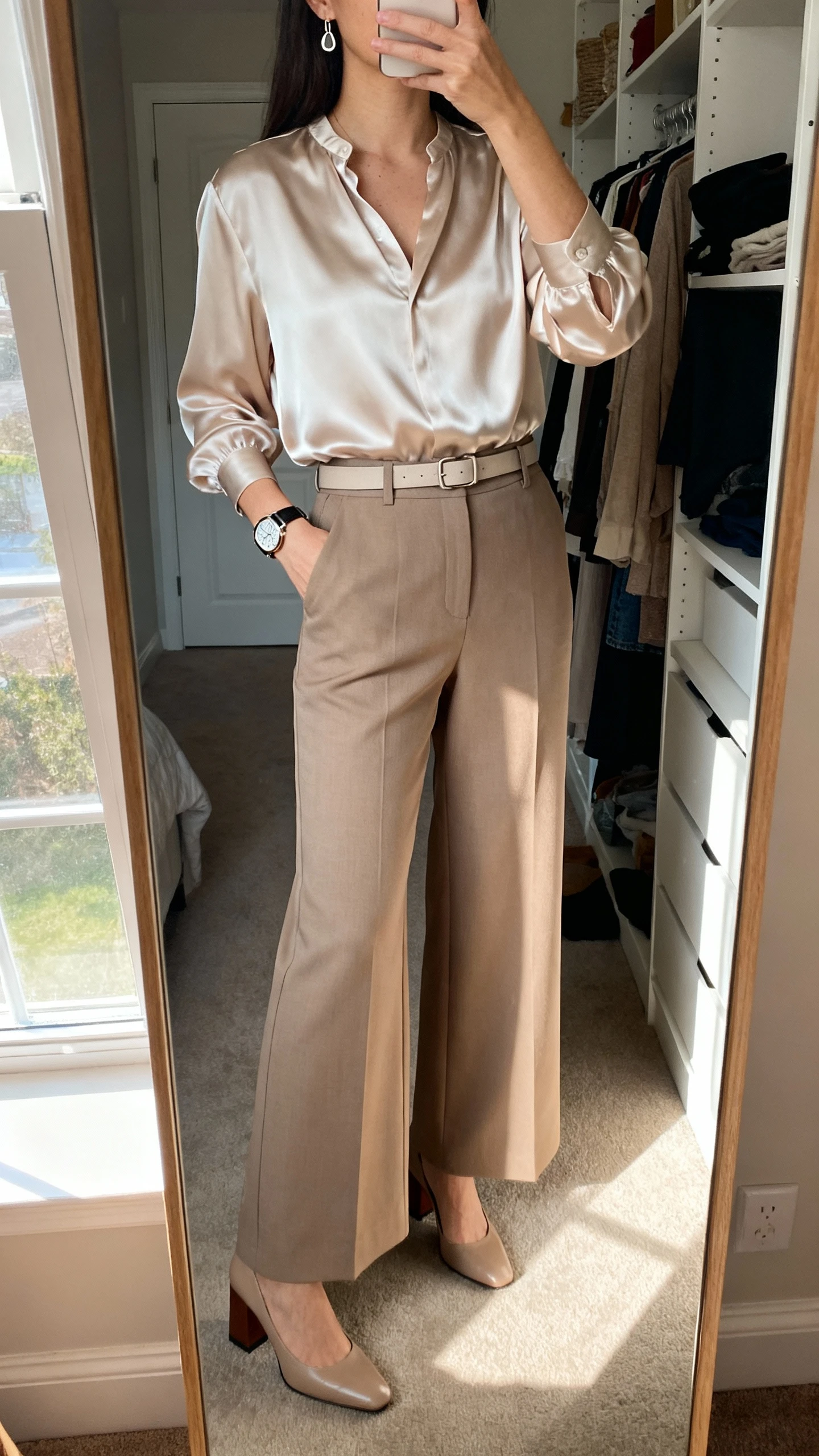 iPhone mirror selfie of a woman wearing high-waisted wide-leg tailored trousers with a silky blouse, block-heel pumps, a minimal belt, simple drop earrings, and a structured watch, face not visible, cropped at neck, closet full-length mirror, morning window light, iPhone photo quality.