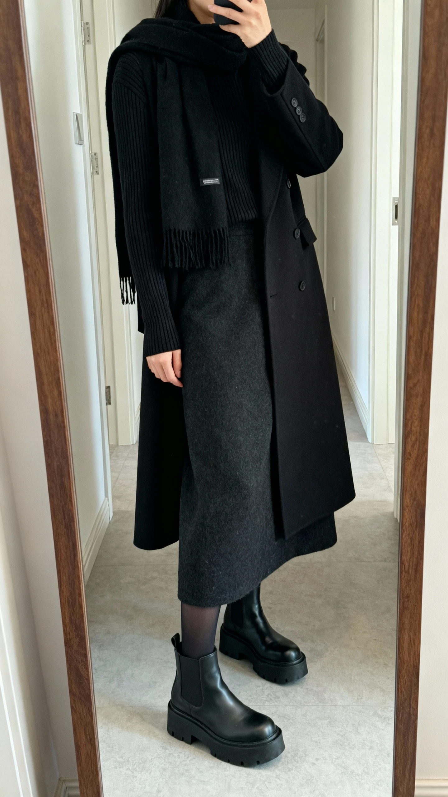 iPhone mirror selfie side angle of a woman in a monochrome all-black look: ribbed knit long-sleeve, wool-blend maxi skirt, long tailored overcoat, opaque tights, platform Chelsea boots, and a black scarf, face not visible, cropped at shoulders, hallway full-length mirror, natural daylight, iPhone photo quality.