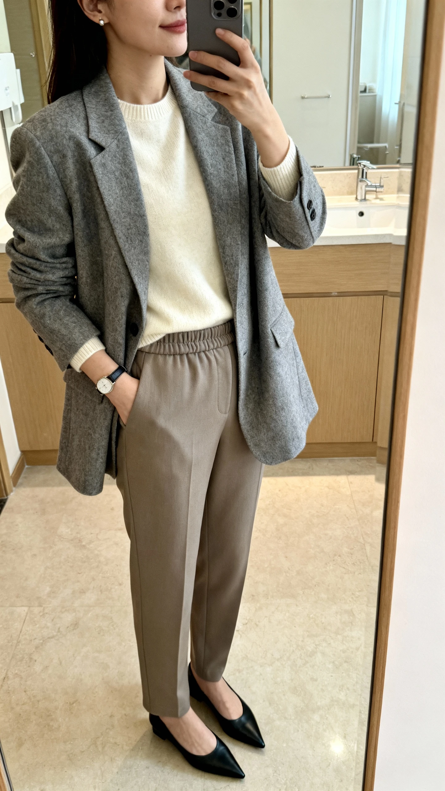 iPhone mirror selfie side angle of a woman in a relaxed-fit heather gray blazer over a cream lightweight cashmere crewneck, tailored straight-leg trousers with a soft waistband, pointed-toe black flats, minimal stud earrings and slim watch, face not visible, bathroom mirror, soft indoor lighting, iPhone photo quality.