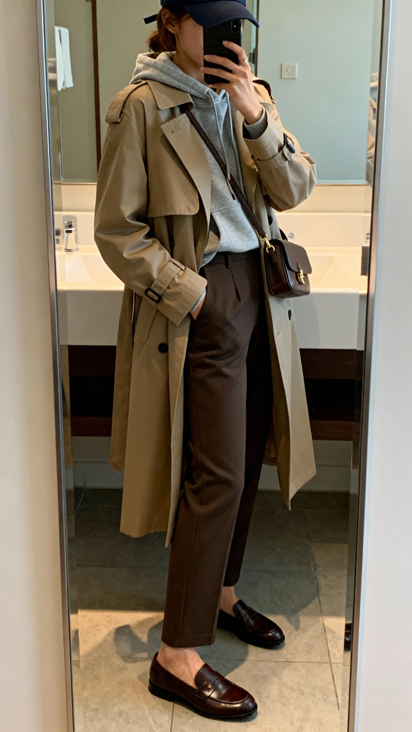 iPhone mirror selfie side angle of a woman wearing a slim hoodie layered under a classic trench coat, straight-leg trousers, sleek leather loafers, small shoulder bag, and a baseball cap, face not visible, bathroom mirror with soft indoor lighting, iPhone photo quality.