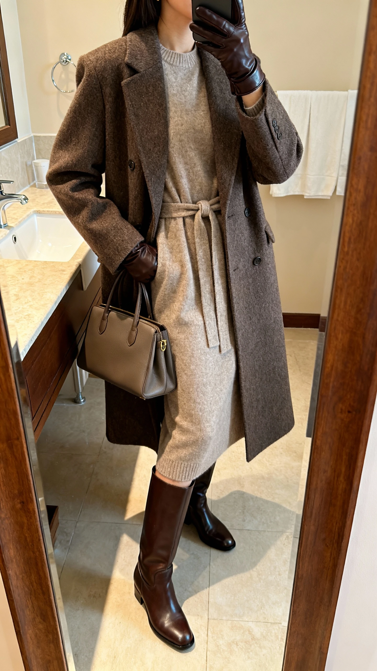 iPhone side-angle mirror selfie of a woman in a belted cashmere long dress layered with a tailored wool coat, sleek knee-high leather boots, leather gloves, and a structured tote, face not visible, bathroom full-length mirror, warm indoor lighting, iPhone photo quality.
