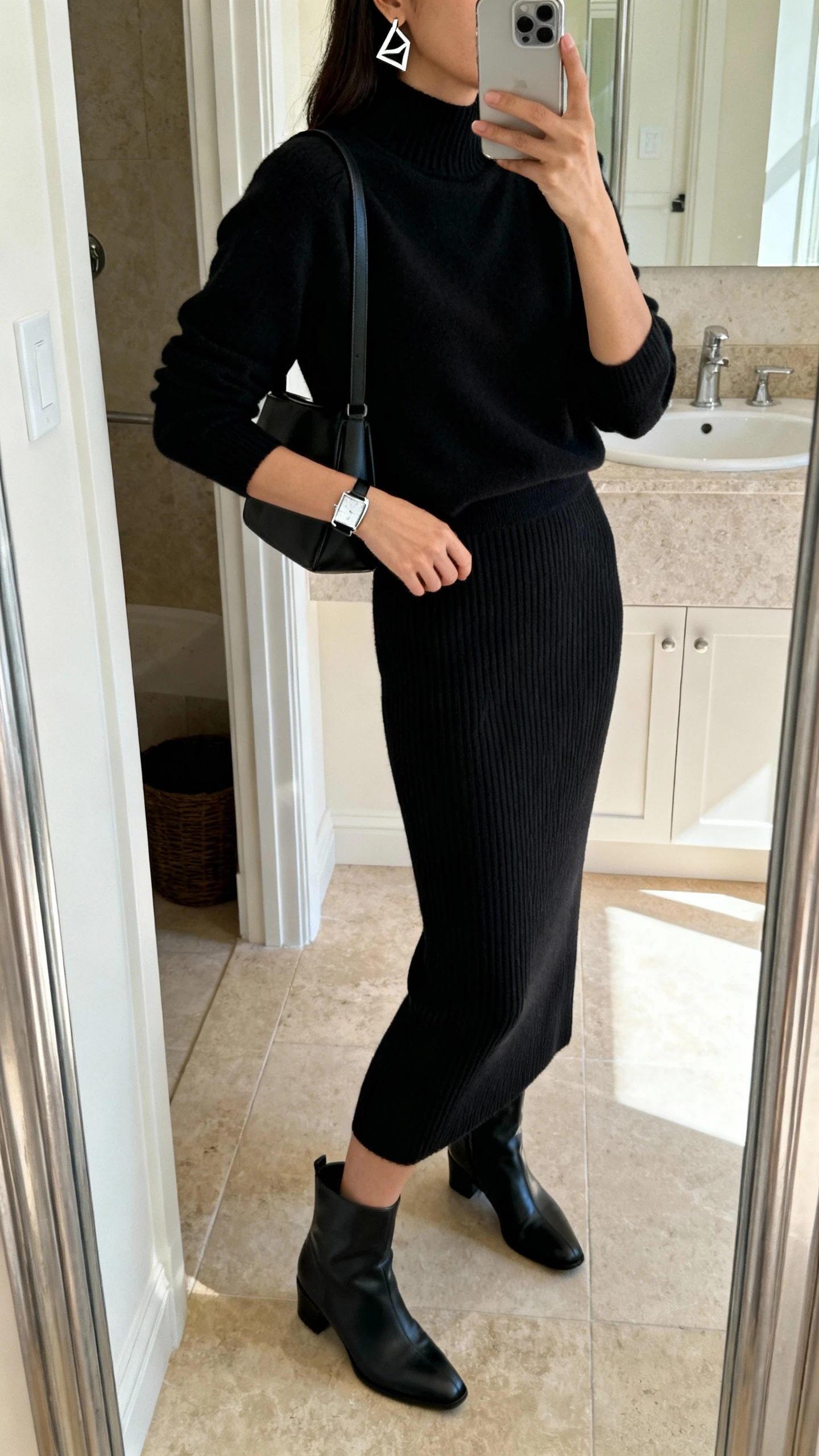 iPhone side-angle mirror selfie of a woman in a black ribbed knit column skirt set with matching mock-neck top, ankle boots, minimal shoulder bag, geometric earrings, and a sleek watch, face not visible, bathroom mirror with natural morning light, iPhone photo quality.