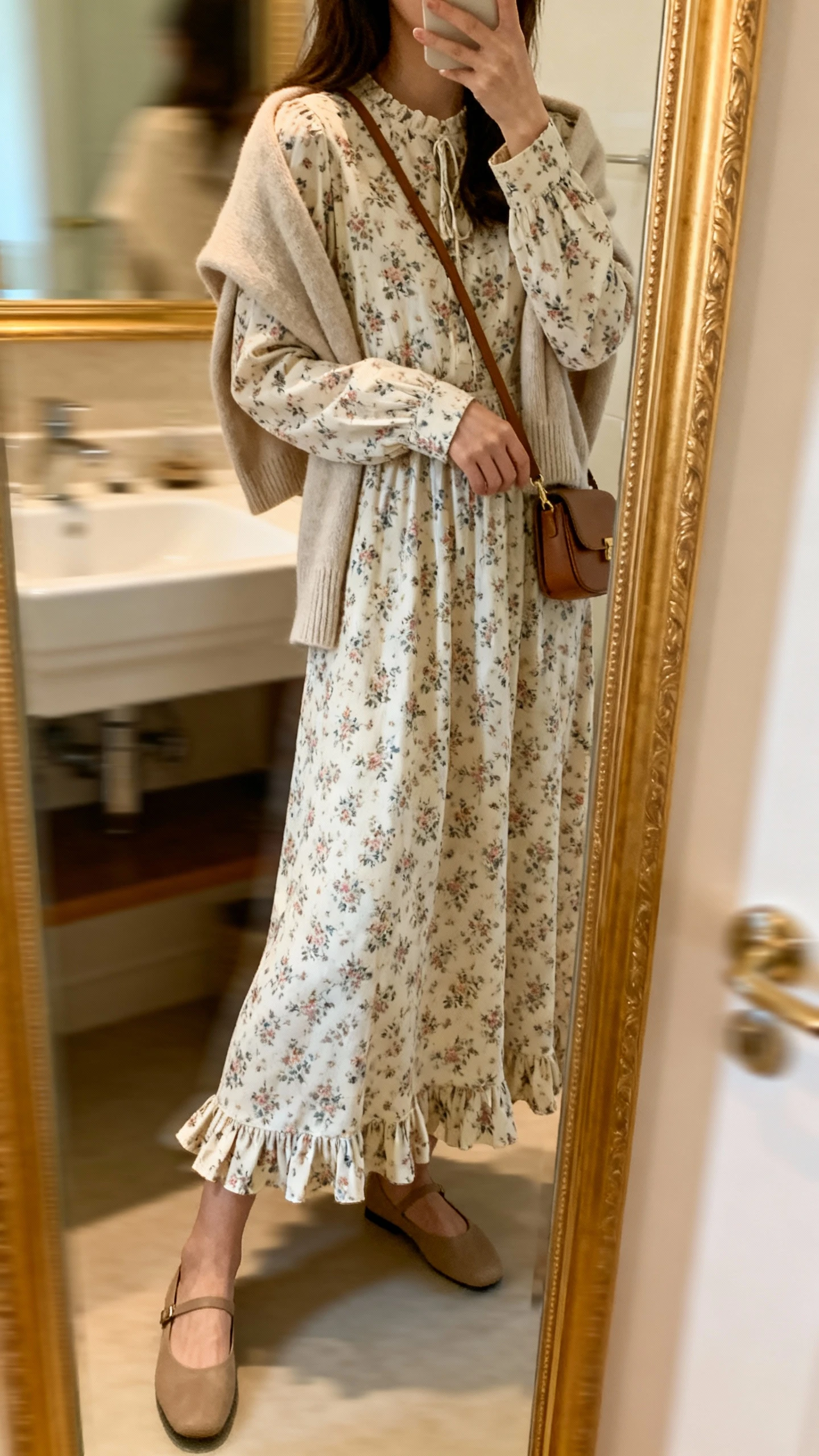 iPhone side-angle mirror selfie of a woman in a cottage-classic floral maxi dress with long sleeves and subtle ruffle hem, soft cardigan over shoulders, small crossbody bag, and simple flats, face not visible, bathroom full-length mirror, indoor natural light, iPhone photo quality.