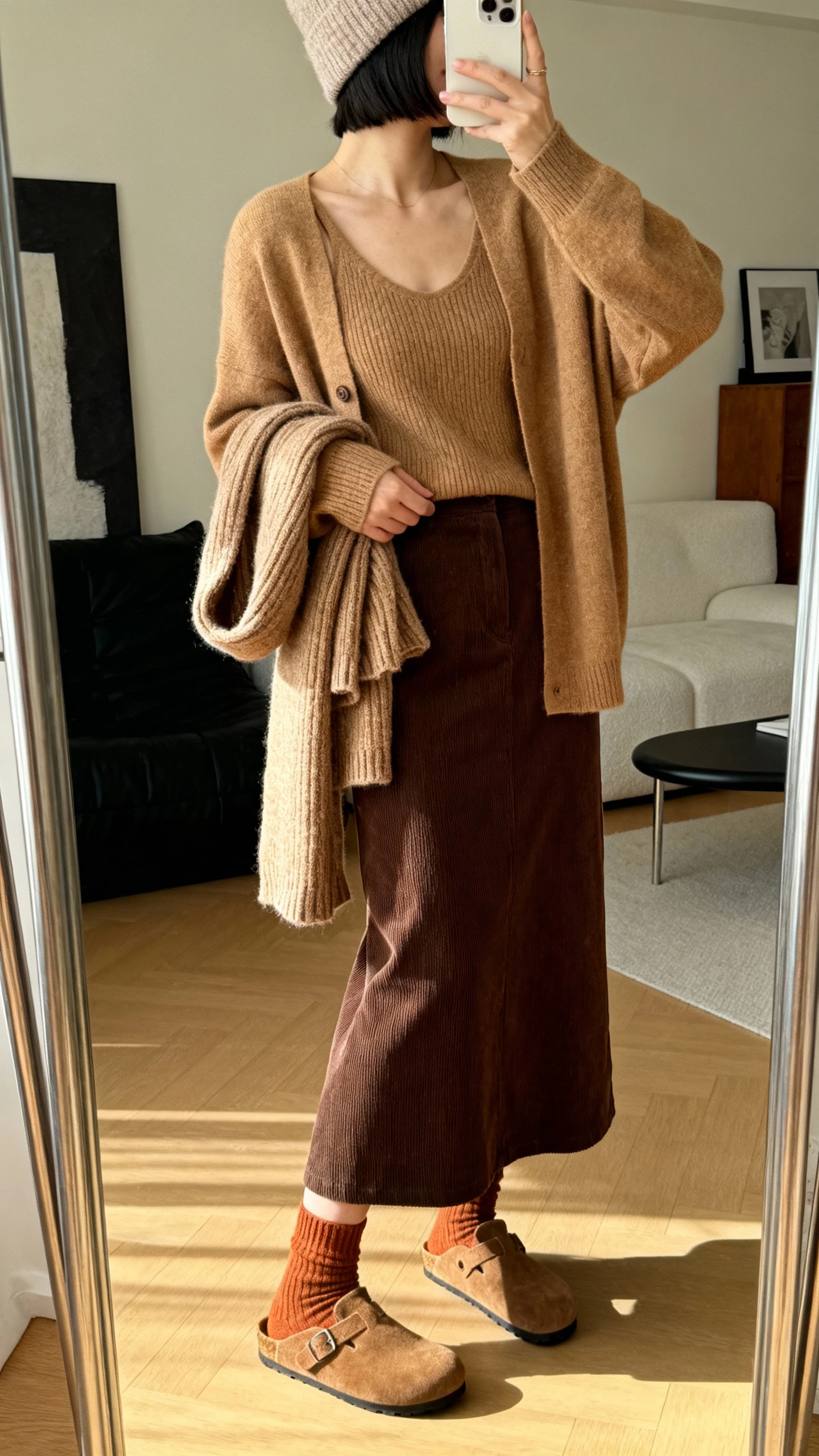iPhone side-angle mirror selfie of a woman in a cozy cardigan set (matching knit tank and cardigan) in tan, paired with a chocolate corduroy maxi skirt, warm socks with suede clogs, and a soft wool beanie, holding a slouchy knit scarf, face not visible, living room mirror, warm indoor lighting, iPhone photo quality.