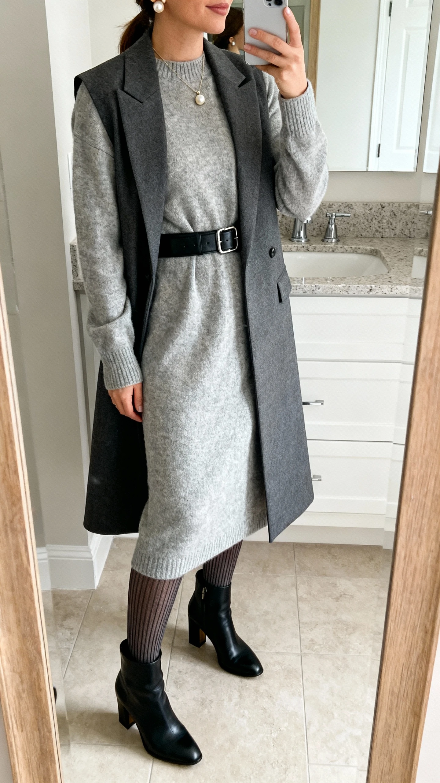 iPhone side-angle mirror selfie of a woman in a heather gray midi sweater-dress cinched with a black leather belt, layered with a longline tailored vest, black heeled ankle boots, ribbed tights, pearl studs, and a delicate pendant necklace; face not visible, bathroom mirror, soft indoor lighting, iPhone photo quality.