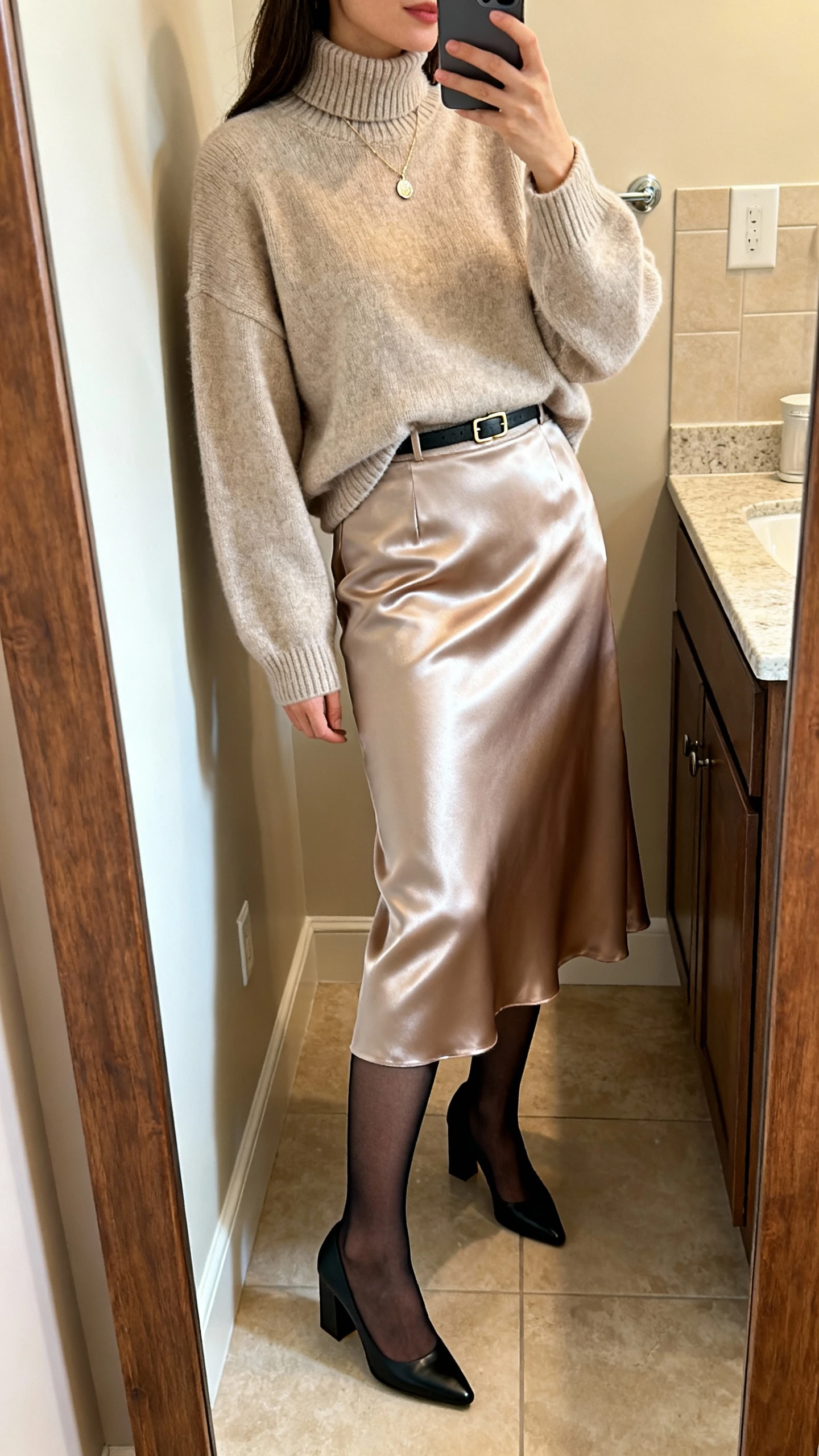 iPhone side‑angle mirror selfie of a woman wearing a satin midi skirt with a fine knit turtleneck layered under a relaxed crewneck sweater, sheer black tights, block‑heel pumps, delicate pendant necklace, and a slim belt; face not visible, bathroom full‑length mirror, warm indoor lighting, iPhone photo quality.