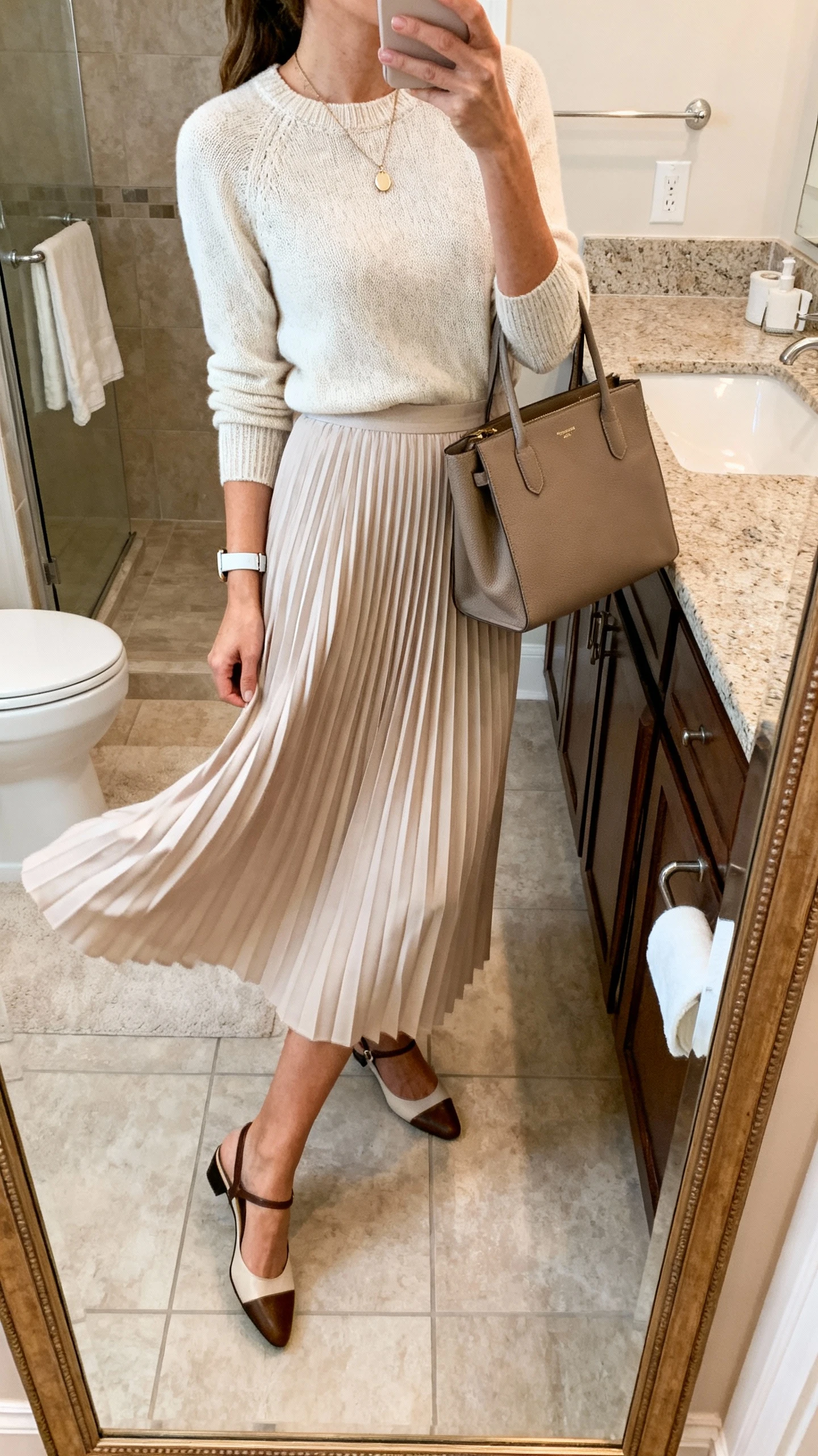 iPhone side-angle mirror selfie of a woman wearing a pleated midi skirt that swishes, tucked lightweight knit top, delicate pendant necklace, slim watch, structured tote, and slingback flats, face not visible, bathroom full-length mirror, soft indoor lighting, iPhone photo quality