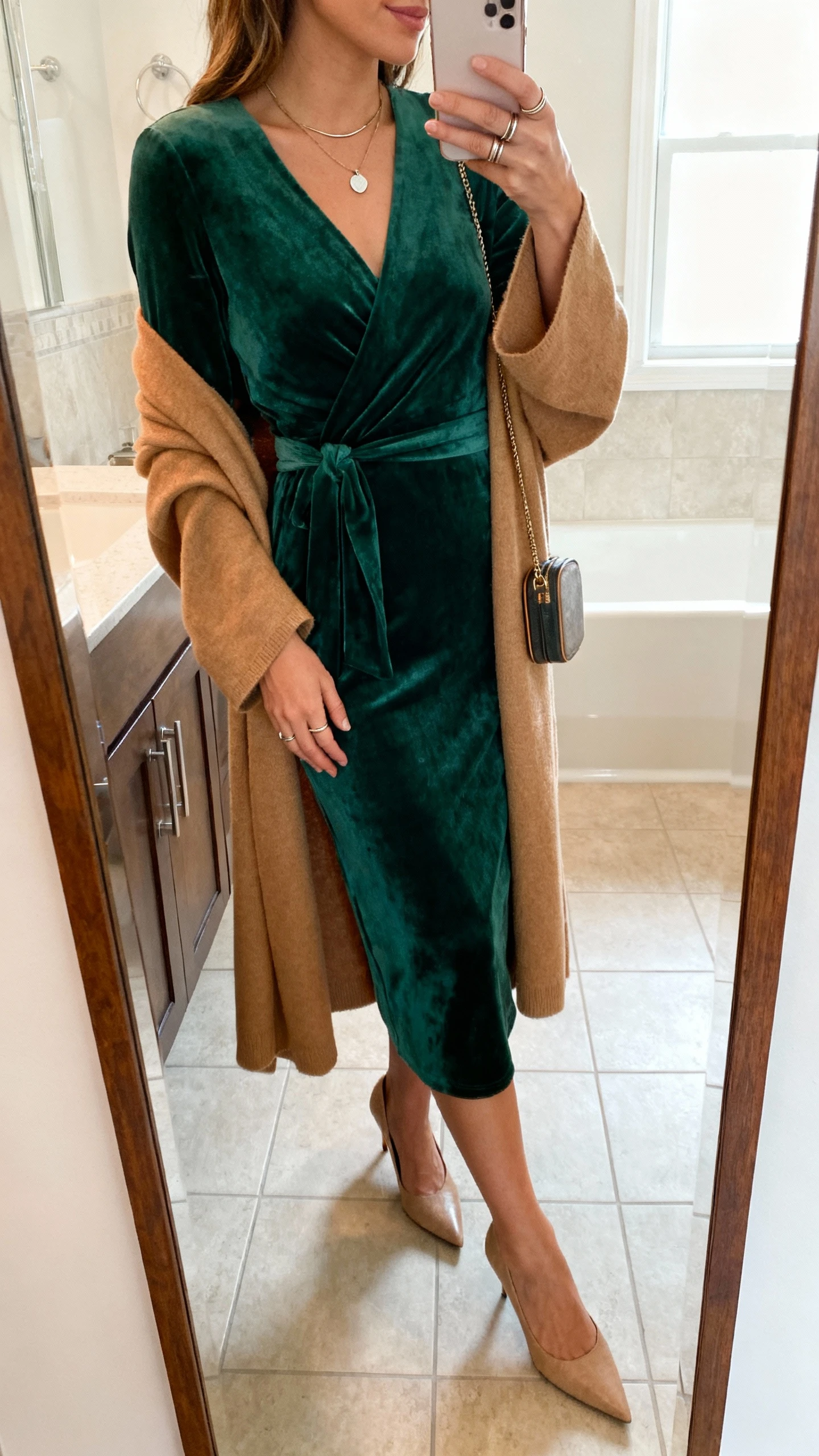 iPhone side-angle mirror selfie of a woman wearing a deep green faux-wrap midi dress with a camel longline duster, pointed kitten heels, a simple pendant necklace, thin stacking rings, and a compact crossbody, face not visible, bathroom mirror, morning light, iPhone photo quality.