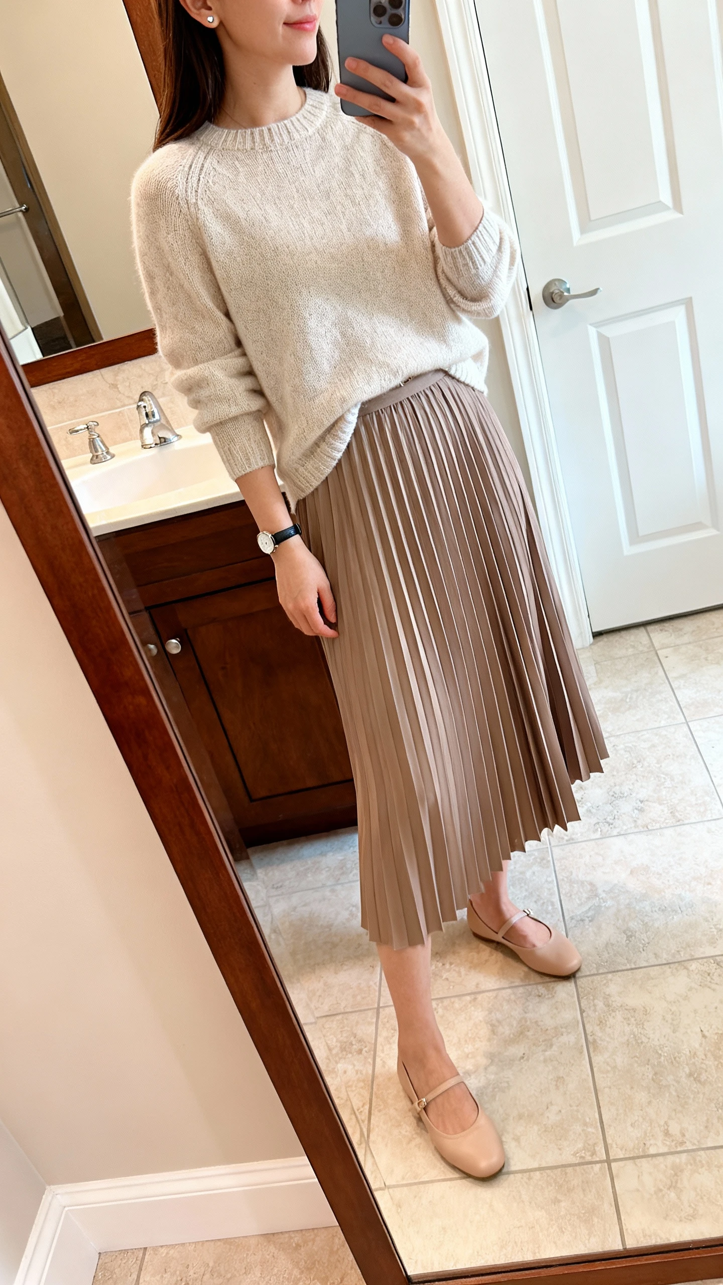 iPhone side-angle mirror selfie of a woman wearing a pleated midi skirt with a soft knit sweater half-tucked, simple stud earrings, slim watch, and ballet flats, face not visible, bathroom full-length mirror, natural indoor lighting, iPhone photo quality.