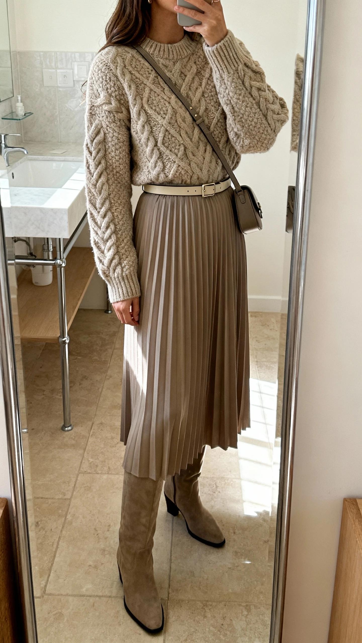 iPhone side-angle mirror selfie of a woman wearing a chunky cable-knit sweater tucked into a pleated midi skirt with knee-high boots and a slim belt, small crossbody, face not visible, bathroom full-length mirror, natural indoor light, iPhone photo quality