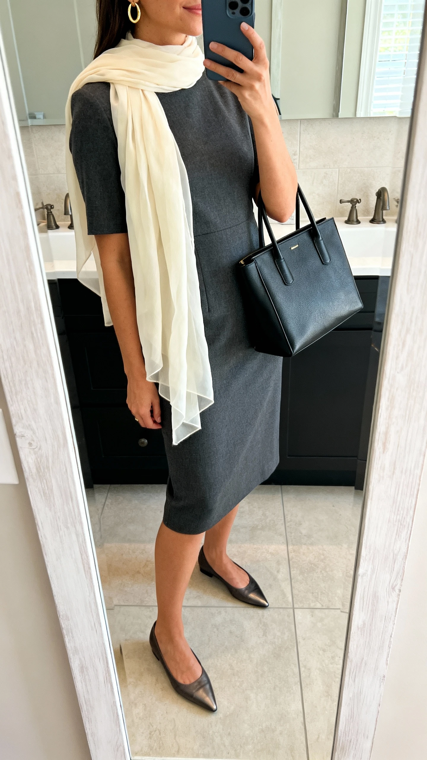 iPhone side-angle mirror selfie of a woman wearing a tailored knee-length sheath dress in charcoal with a lightweight cream chiffon scarf draped over the shoulders, structured black tote, simple gold hoop earrings, and pointed-toe flats, face not visible, bathroom mirror, indoor soft lighting, iPhone photo quality.