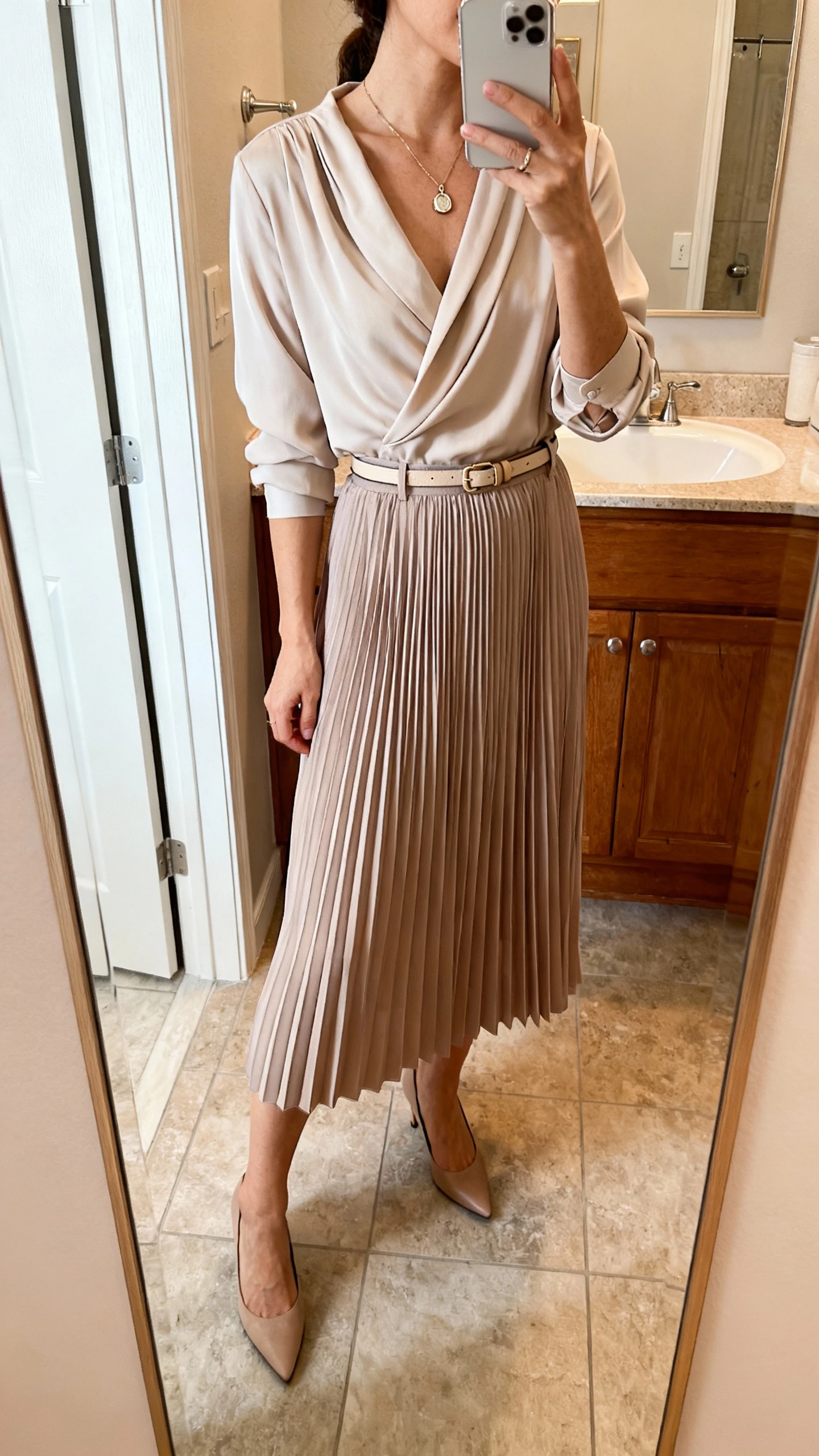 iPhone side-angle mirror selfie of a woman wearing a pleated midi skirt and a soft drapey blouse tucked in, delicate pendant necklace, slim belt, and classic pumps, face not visible, bathroom full-length mirror, natural indoor lighting, iPhone photo quality.