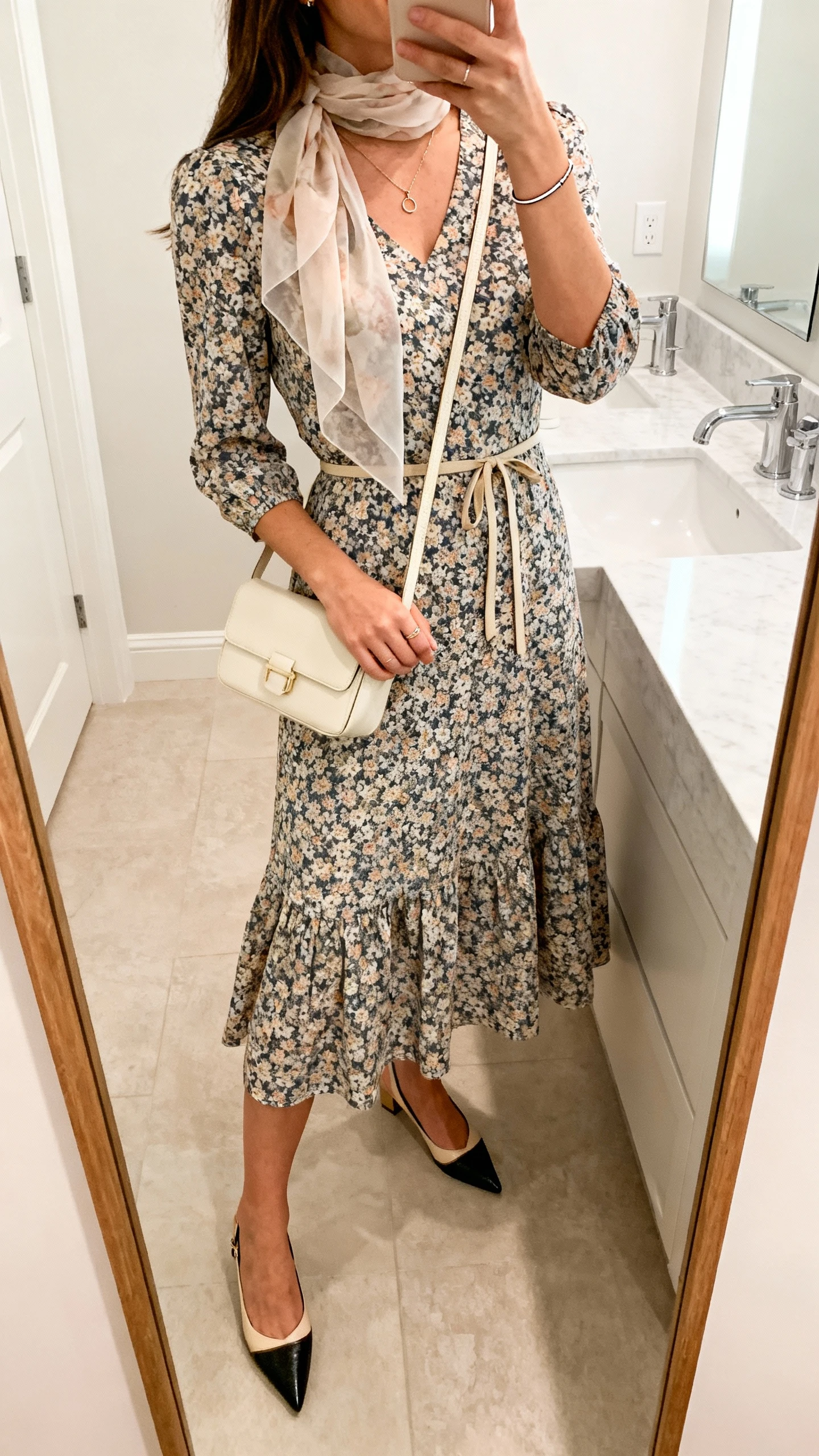iPhone side-angle mirror selfie of a woman wearing a floral A-line dress with a modest neckline and 3/4 sleeves, a lightweight chiffon scarf, pointed-toe slingbacks, a dainty pendant necklace with a thin bracelet, a small cream crossbody bag, and a thin waist tie, face not visible, bathroom full-length mirror, soft indoor lighting, iPhone photo quality.