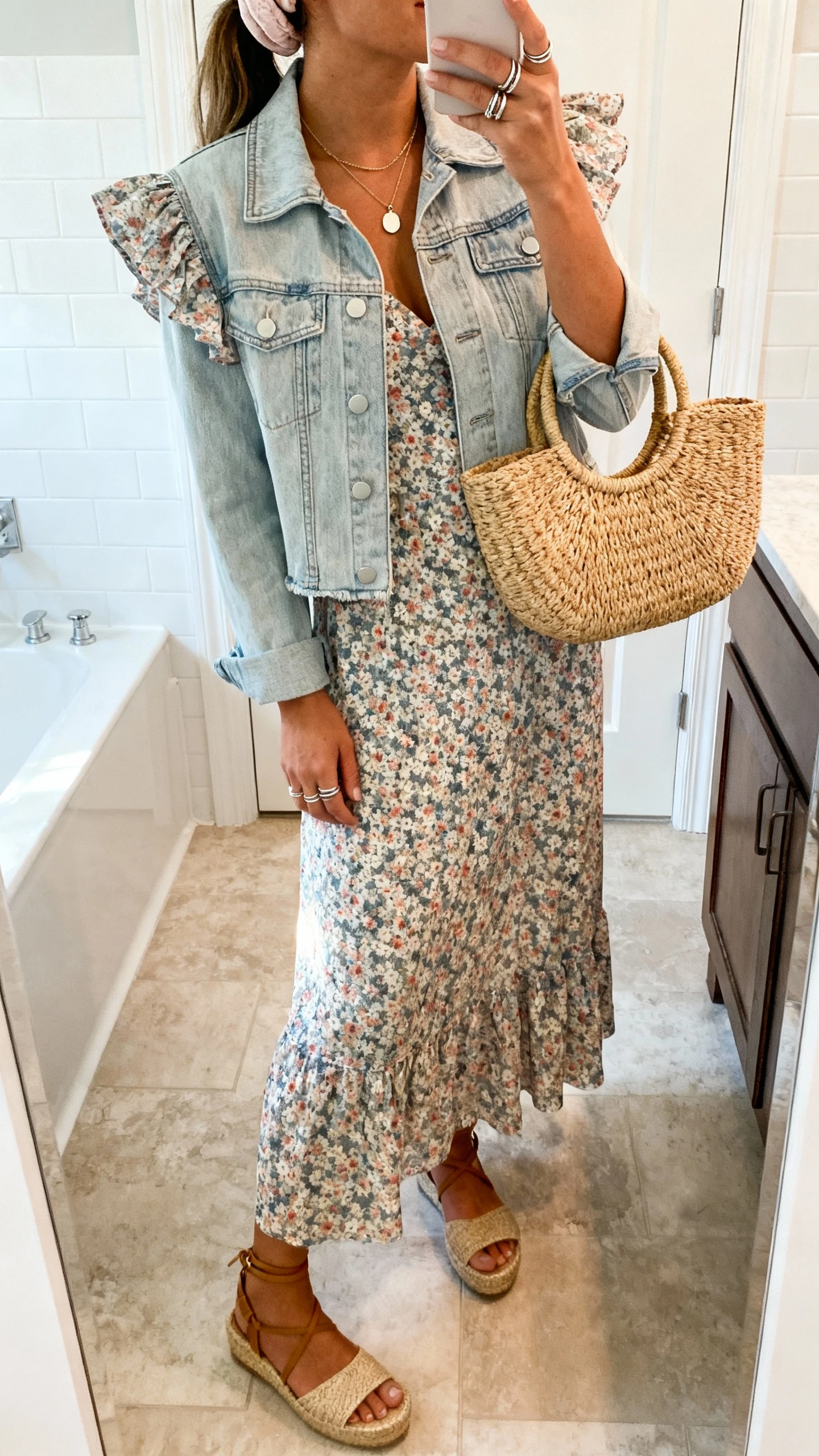 iPhone side-angle mirror selfie of a woman wearing a flutter-sleeve floral maxi dress with a light-wash cropped denim jacket, espadrille wedges, a straw top-handle bag, a simple pendant necklace, stackable rings, and a soft headband; face not visible, bathroom mirror, morning light, iPhone photo quality.