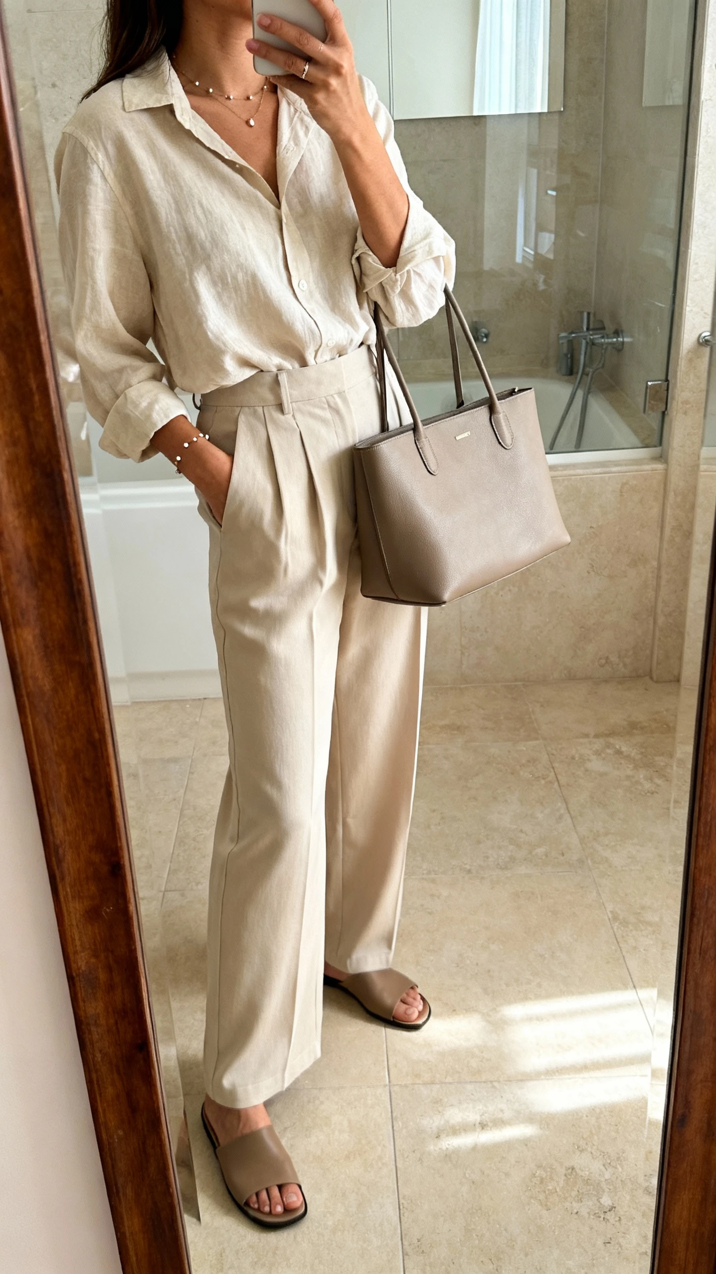 iPhone side-angle mirror selfie of a woman wearing a soft blouse tucked into wide-leg trousers, delicate studs, flat mules, and a structured tote, face not visible, cropped at shoulders, bathroom mirror, morning indoor lighting, iPhone photo quality.