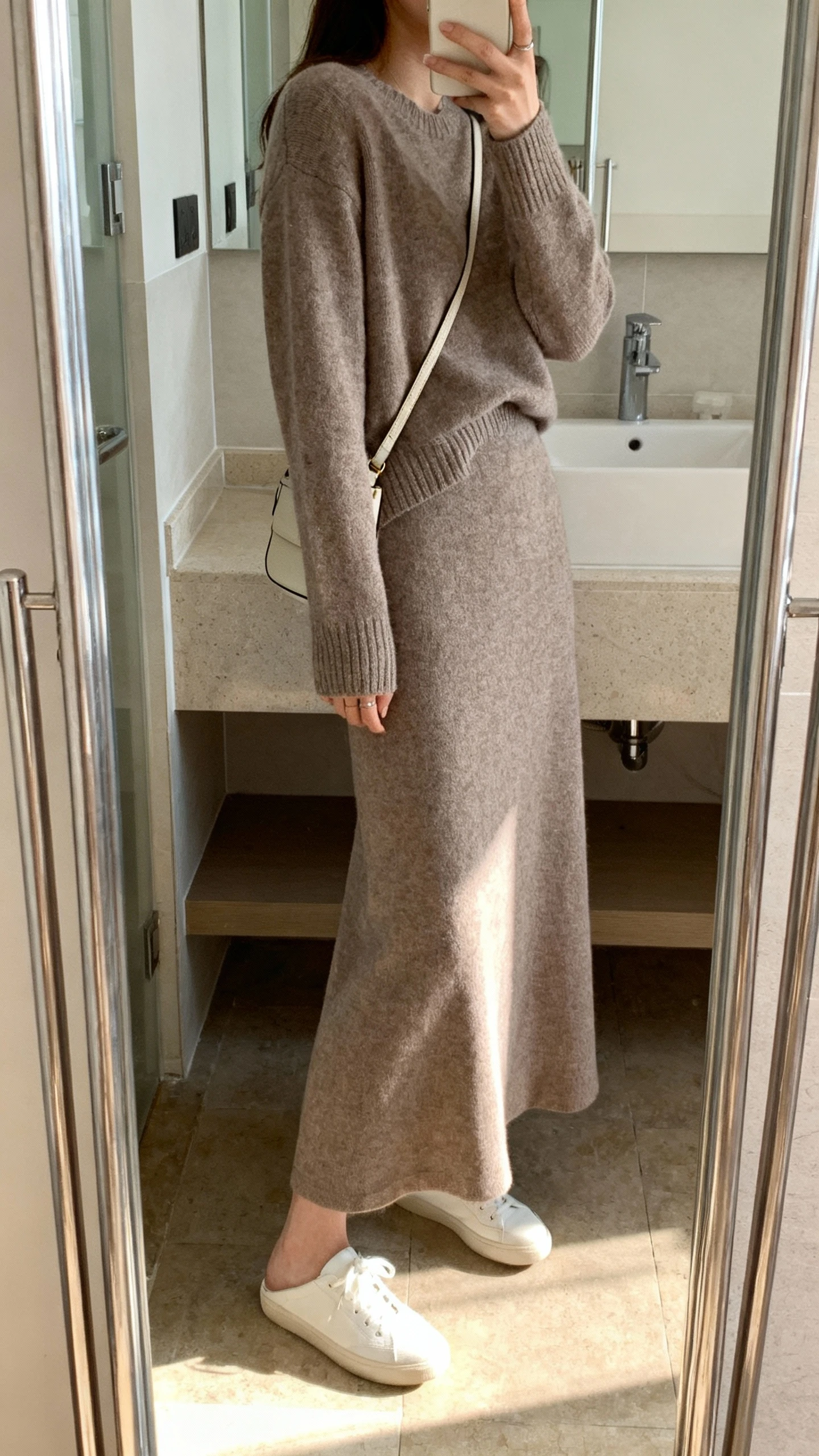iPhone side-angle mirror selfie of a woman wearing a soft knit matching set: long-sleeve sweater and flowy maxi skirt in a muted taupe, paired with minimalist white sneakers and a small crossbody bag, face not visible, bathroom mirror, morning light, iPhone photo quality.