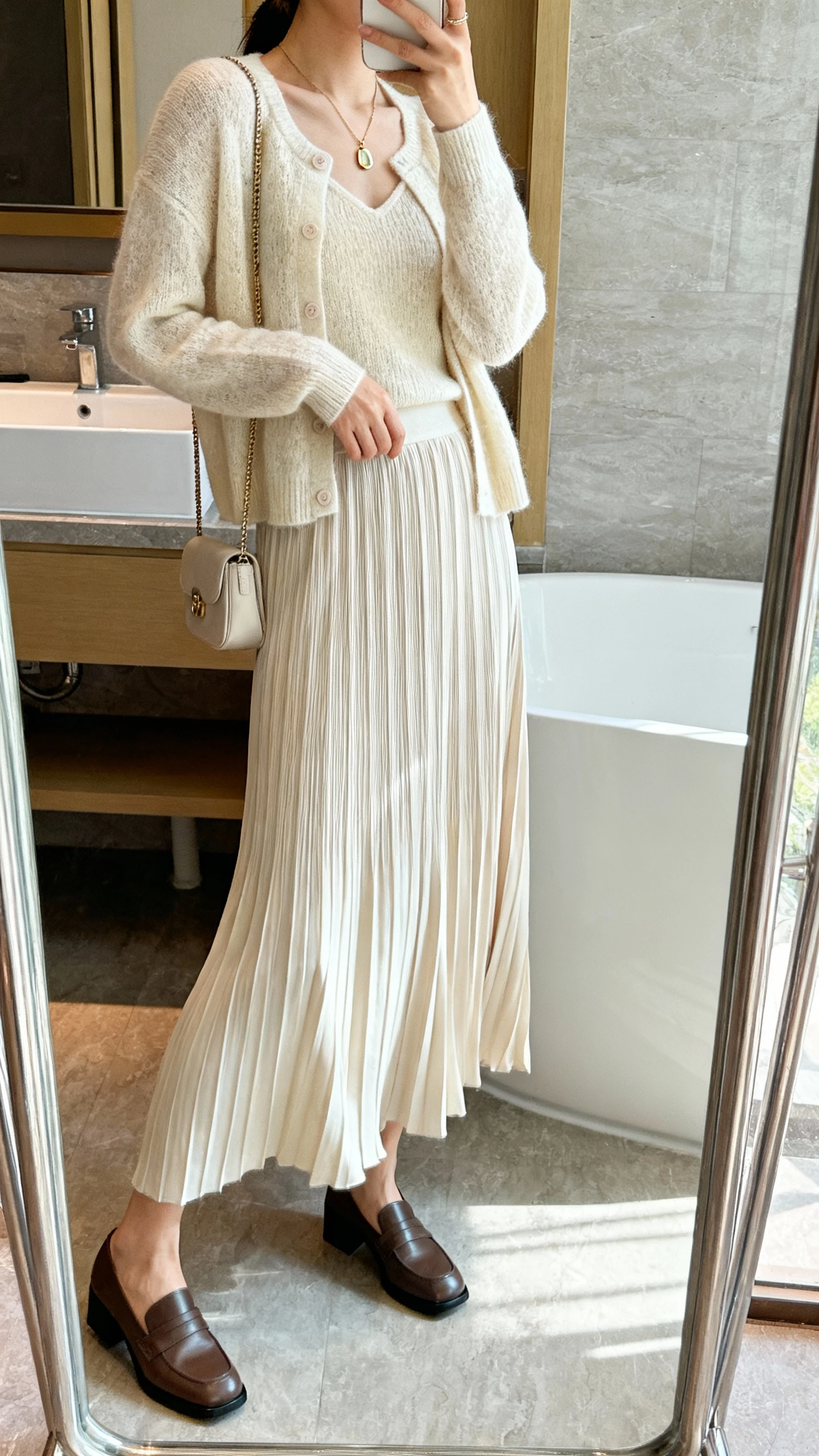 iPhone side-angle mirror selfie of a woman wearing a soft knit cardigan set (matching shell and cardigan) with a pleated maxi skirt that sways, paired with low block-heel loafers, delicate pendant necklace, and a small crossbody, face not visible, bathroom full-length mirror, indoor natural light, iPhone photo quality.