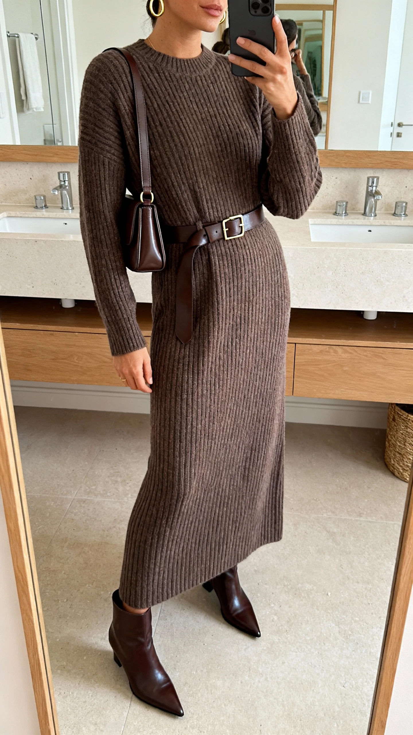 iPhone side-angle mirror selfie of a woman wearing a ribbed belted knit maxi dress, sleek leather belt, minimal gold hoops, slim leather shoulder bag, and pointed leather ankle boots, face not visible, bathroom mirror, soft indoor lighting, iPhone photo quality.